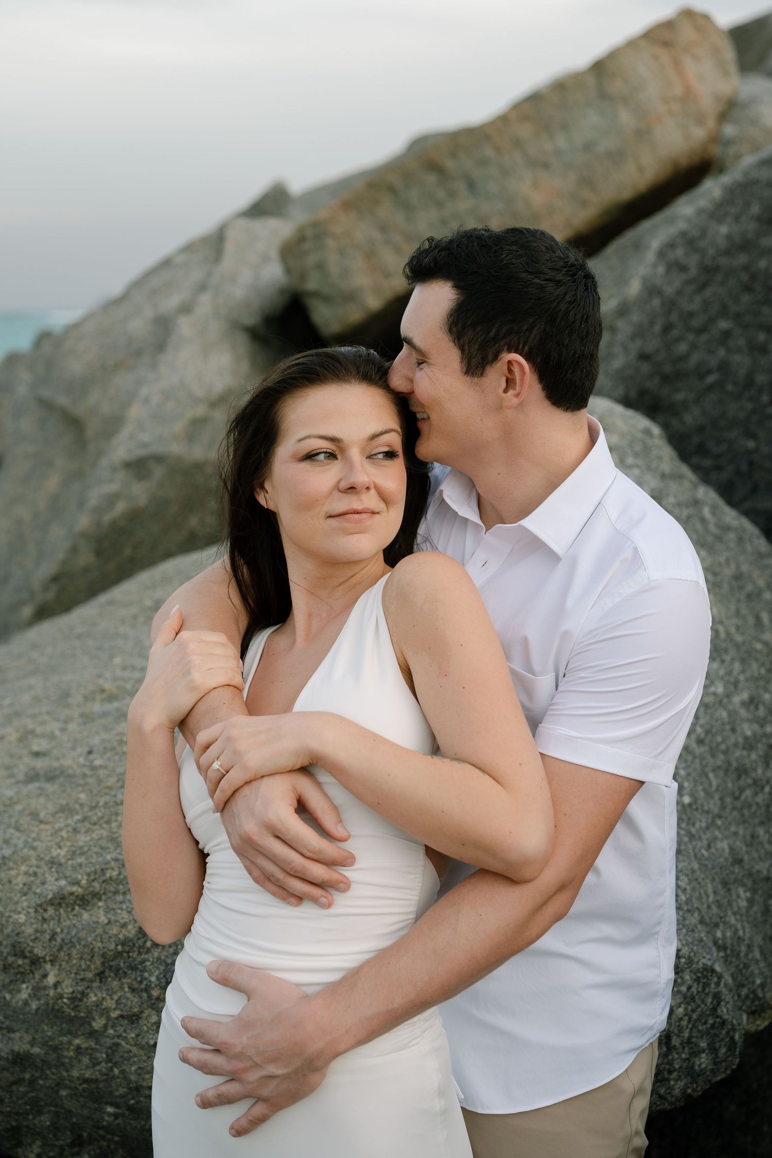 Romantic couple embraces against rocky coastal backdrop in intimate photoshoot series.