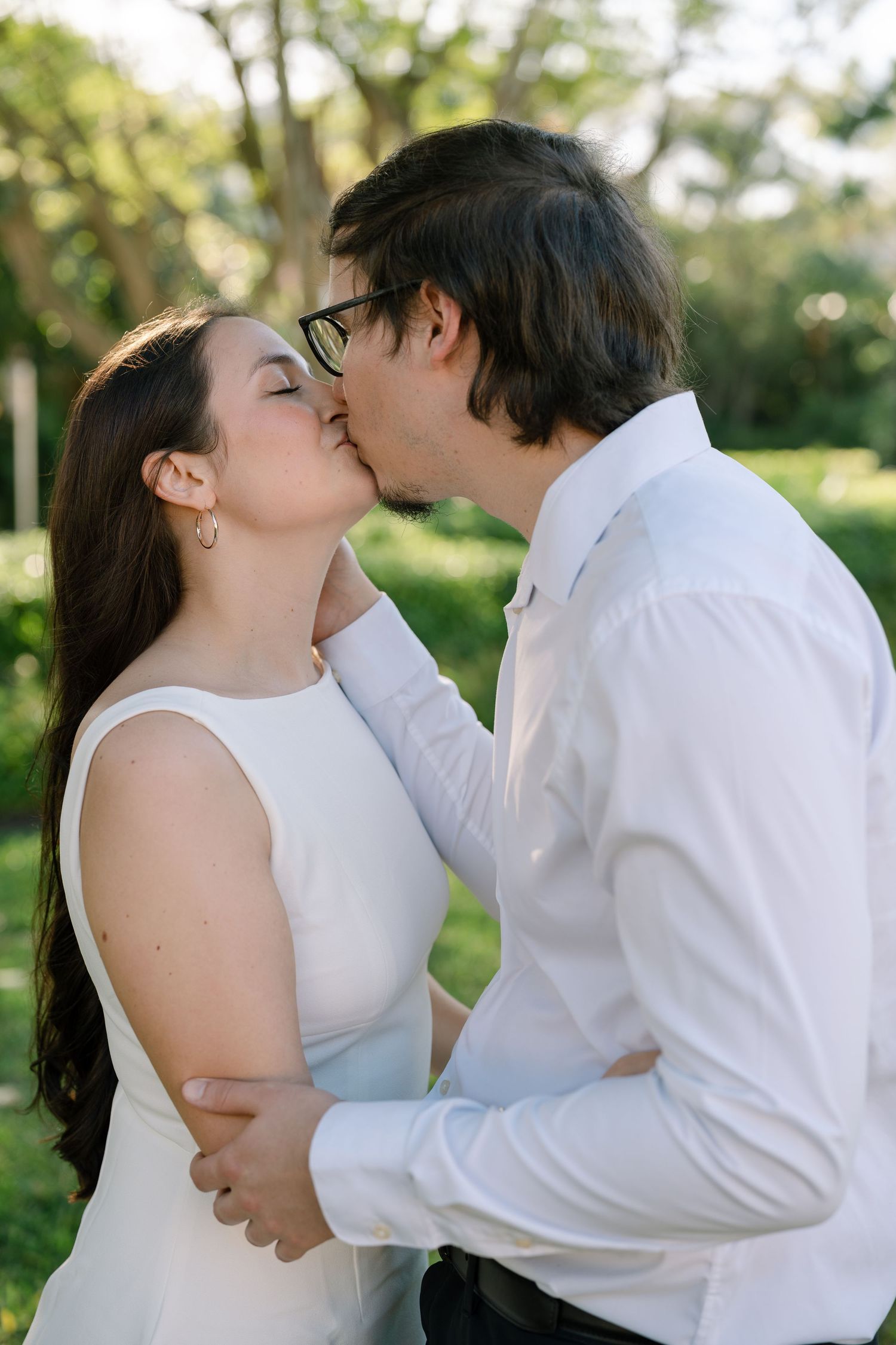 A romantic couple dressed in white shares intimate kisses in a garden setting with soft natural lighting.