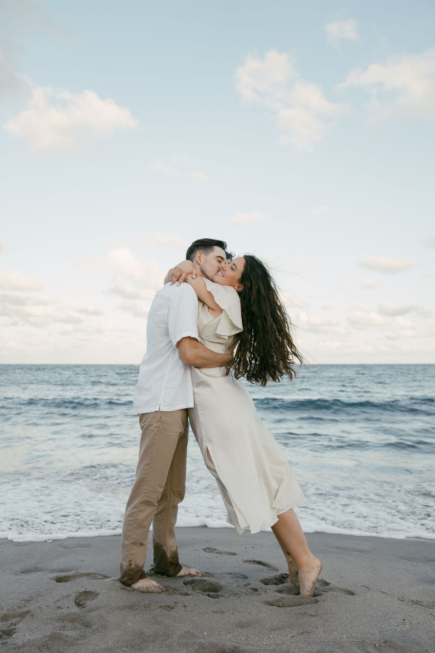 Affectionate couple embraces on a sandy beach at sunset with ocean waves in the background.