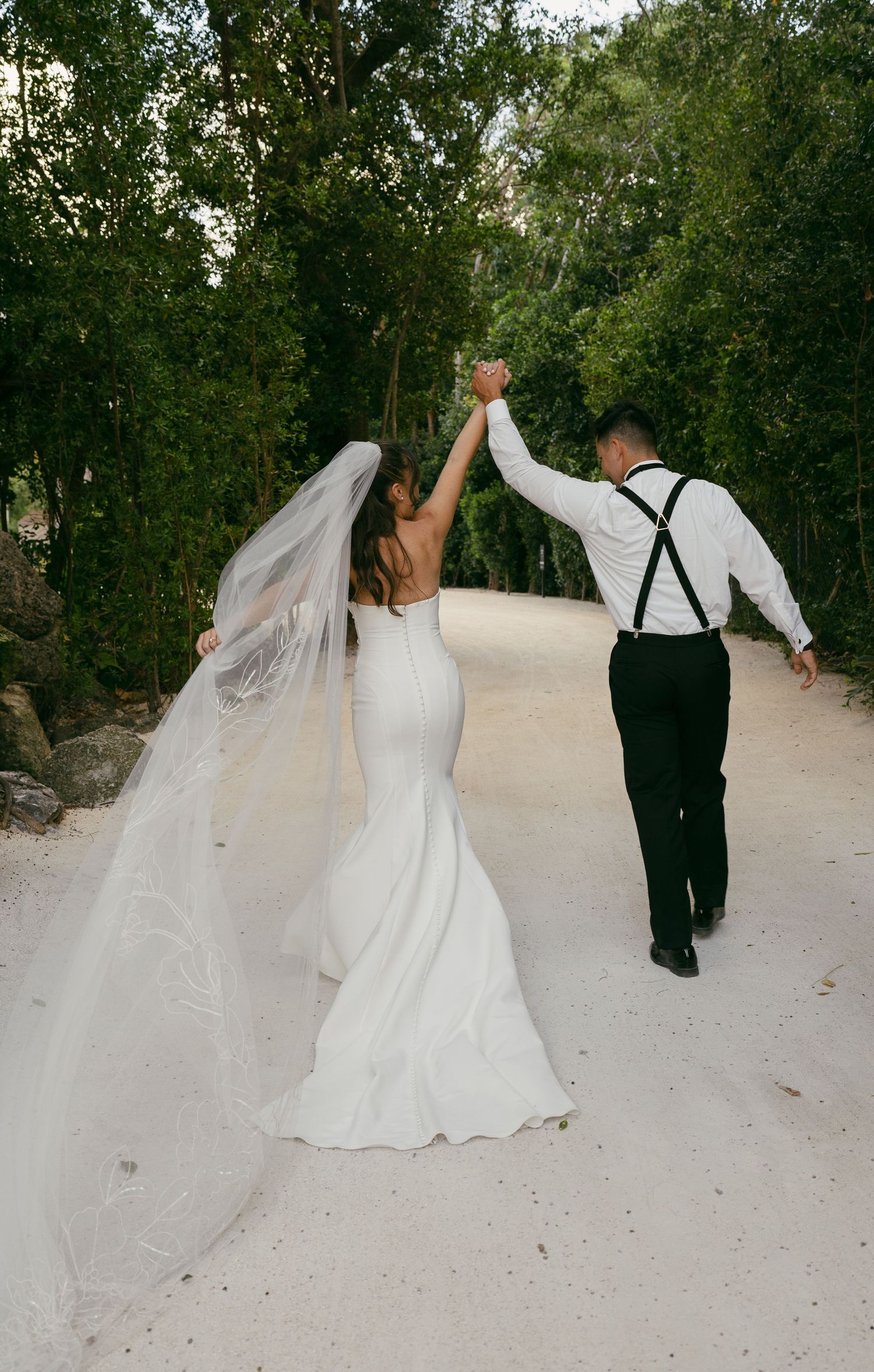 Newlyweds holding hands and walking down a sandy path surrounded by green trees with bride's veil flowing behind.