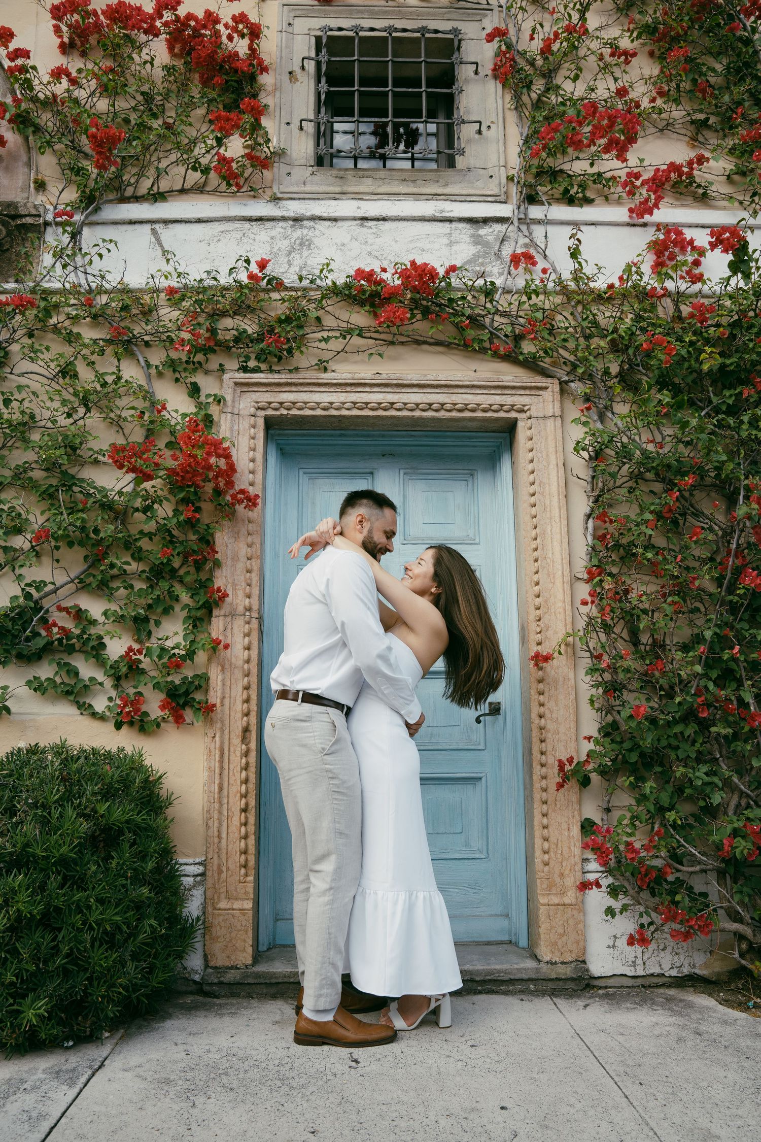 A couple embraces in front of a light blue door framed by climbing red flowers and vines on a white building.