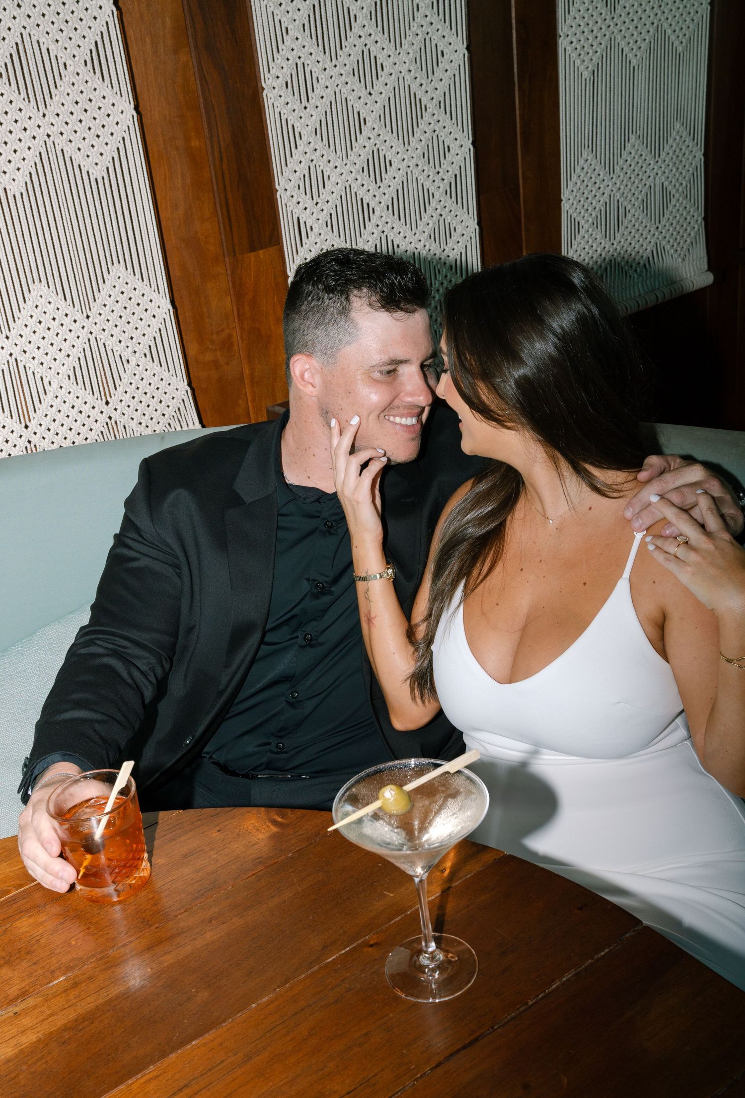 A couple sits at a wooden table in a dimly lit restaurant sharing drinks and intimate conversation.