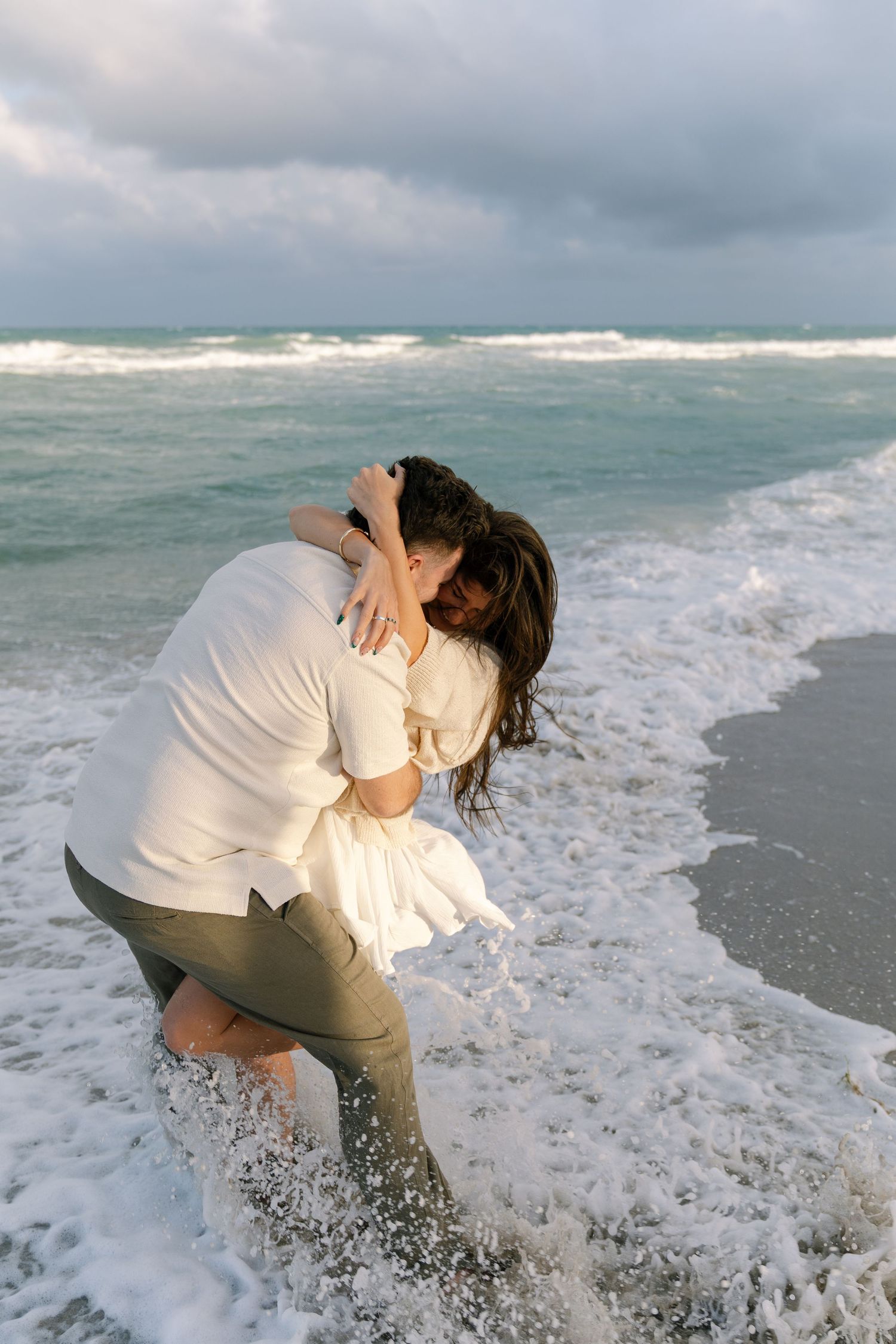A couple embraces in the ocean waves at sunset on a cloudy beach as water splashes around their feet.
