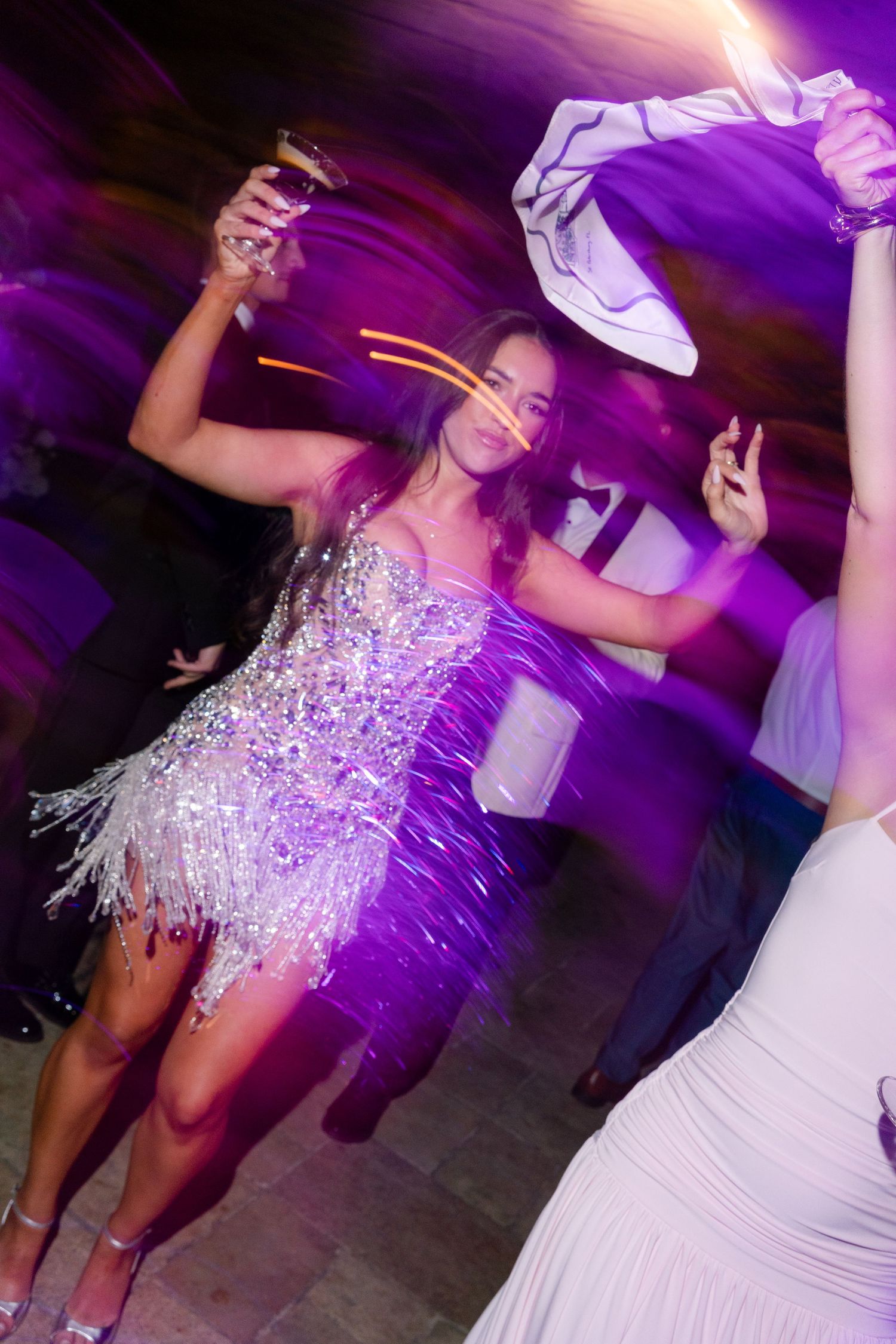 Bride dancing in sparkling silver fringe mini dress under purple lights at wedding reception.