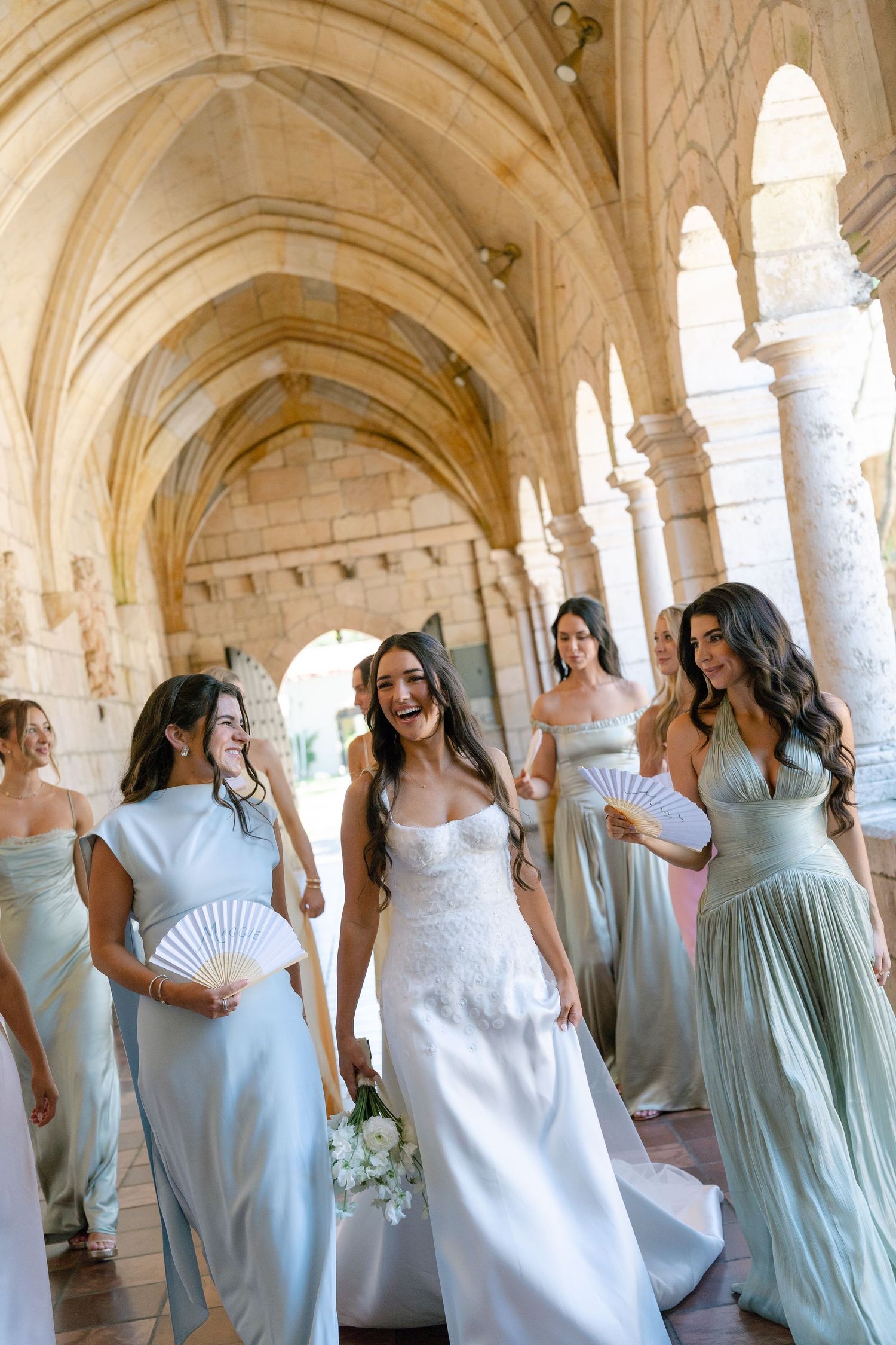 Laughing bride in white gown walks with bridesmaids in sage green dresses through stone archway.