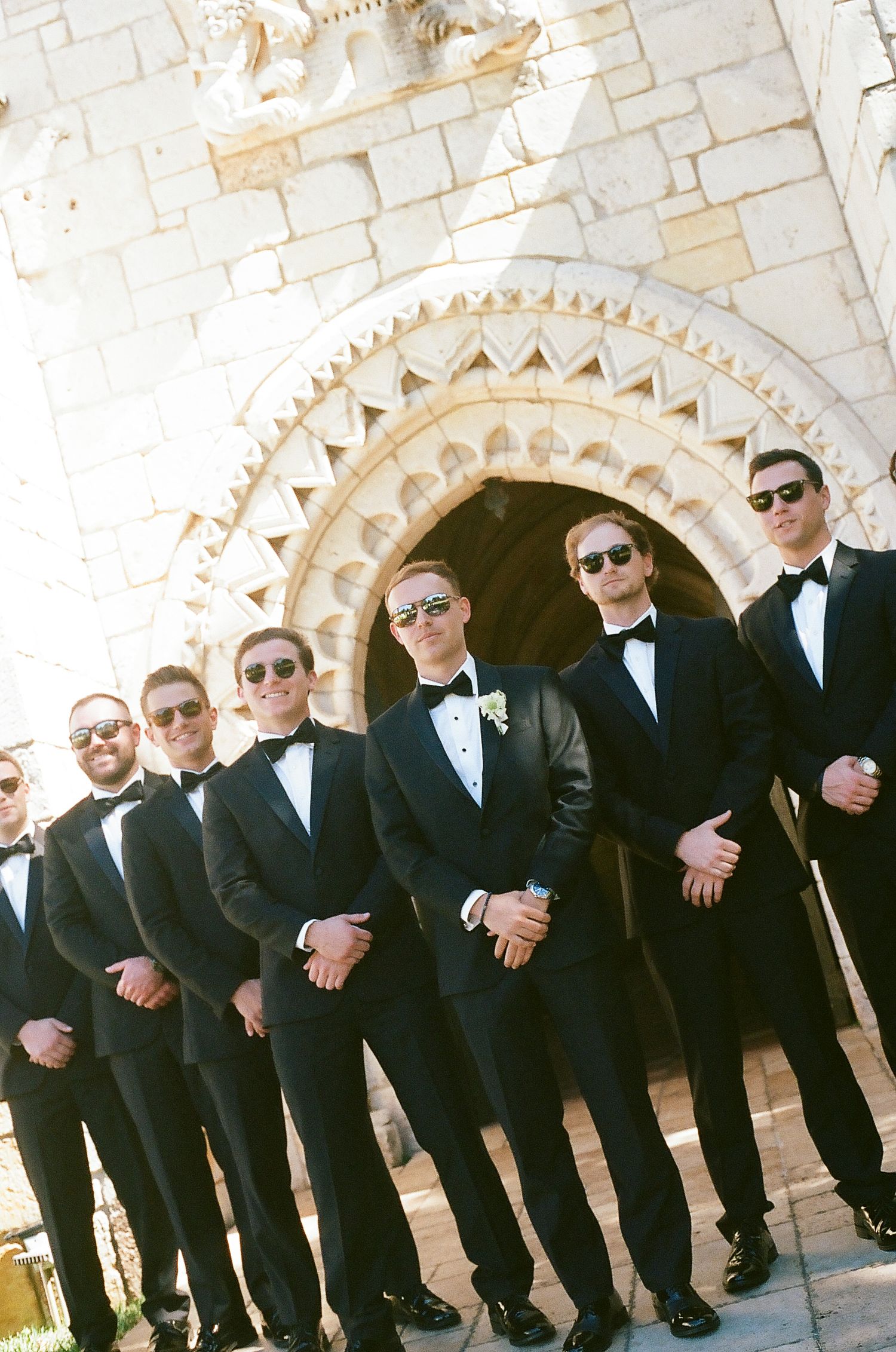 Groom and groomsmen in black tuxedos and sunglasses pose in front of a stone church.