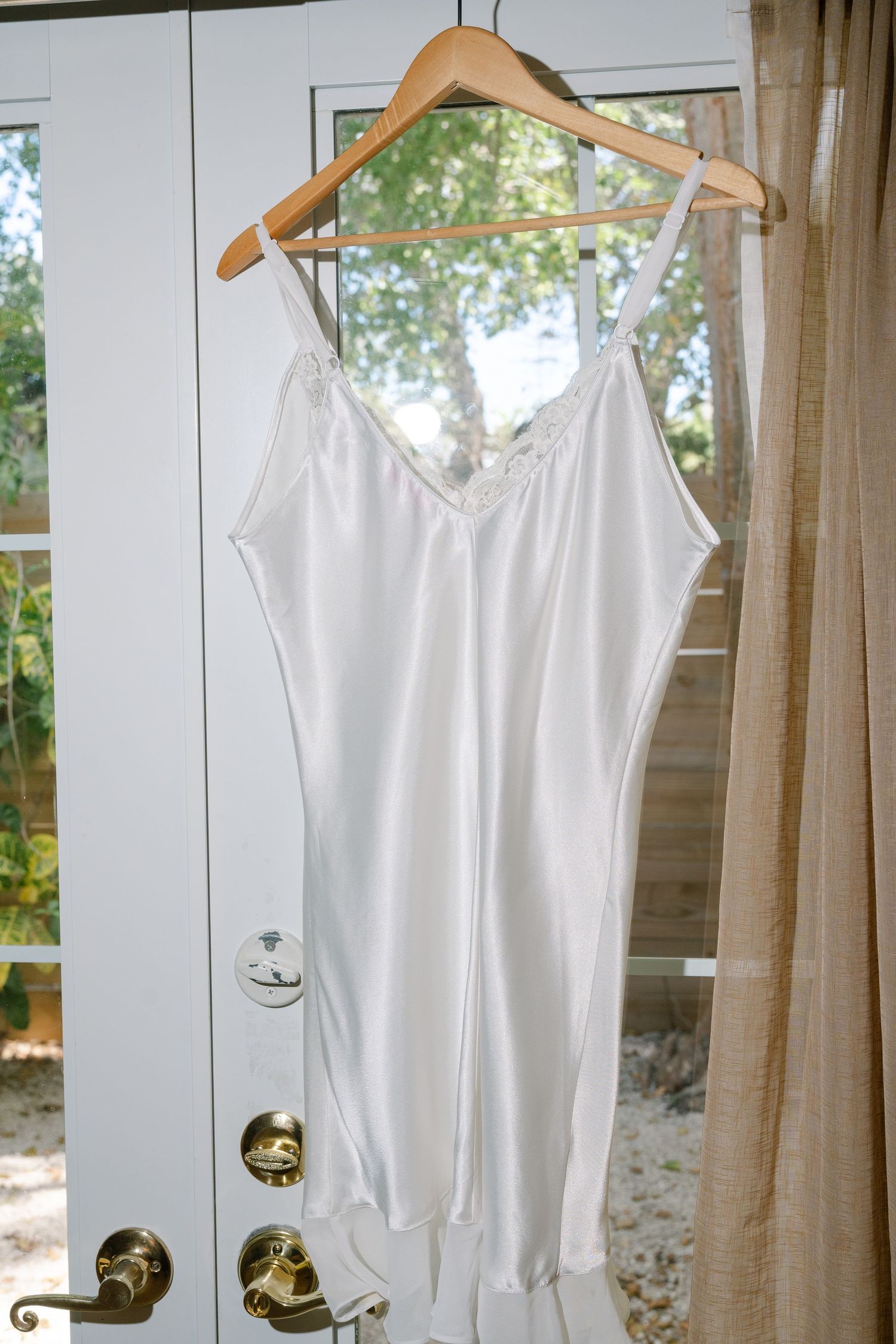 A white lace dress hangs on a wooden hanger against a window.