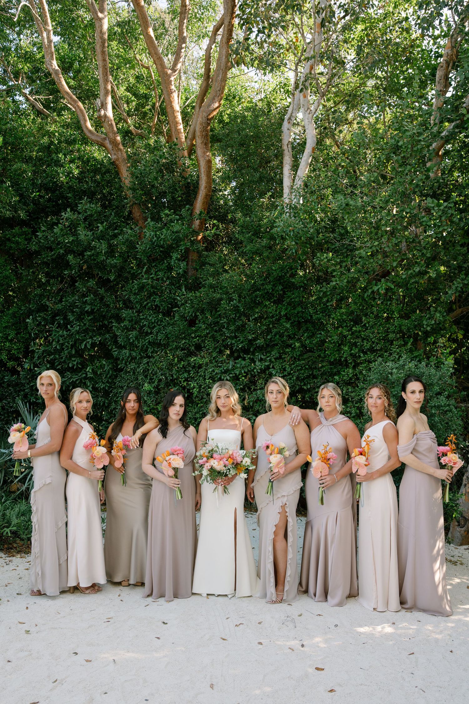 Bridesmaids in elegant neutral dresses pose in a line against lush garden backdrop.