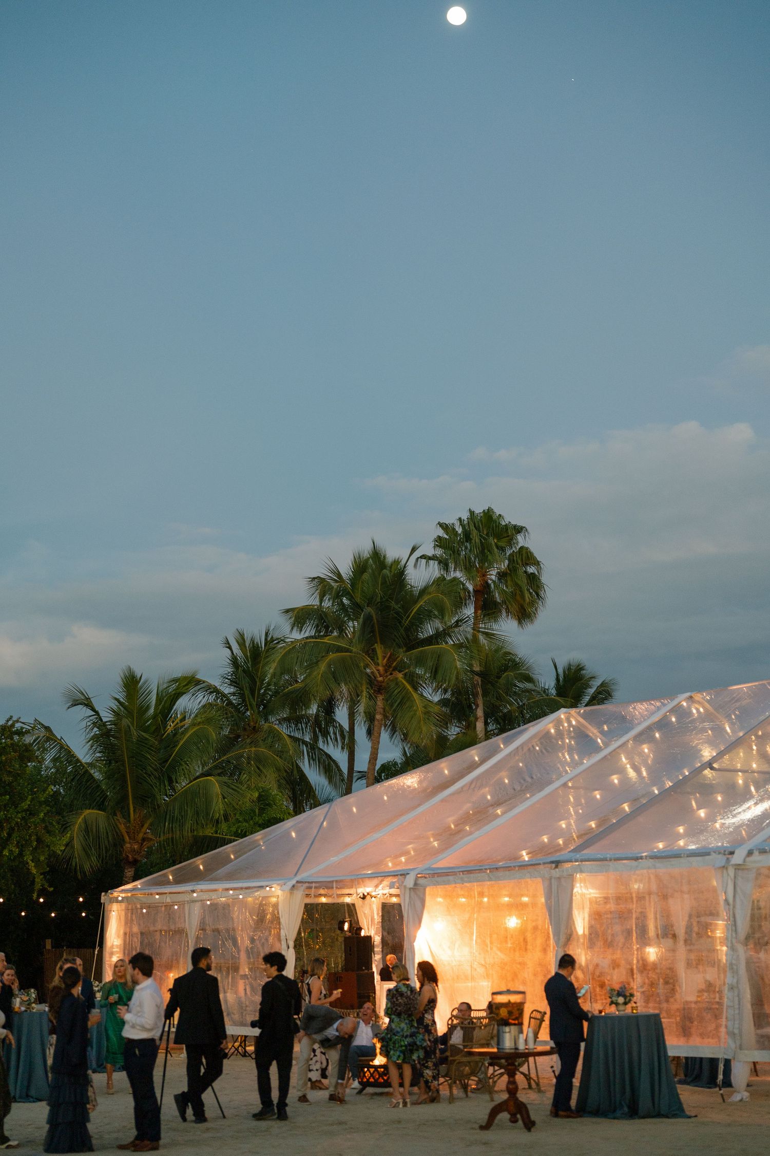 An illuminated clear tent reception venue at dusk with palm trees and a full moon in the tropical evening sky.