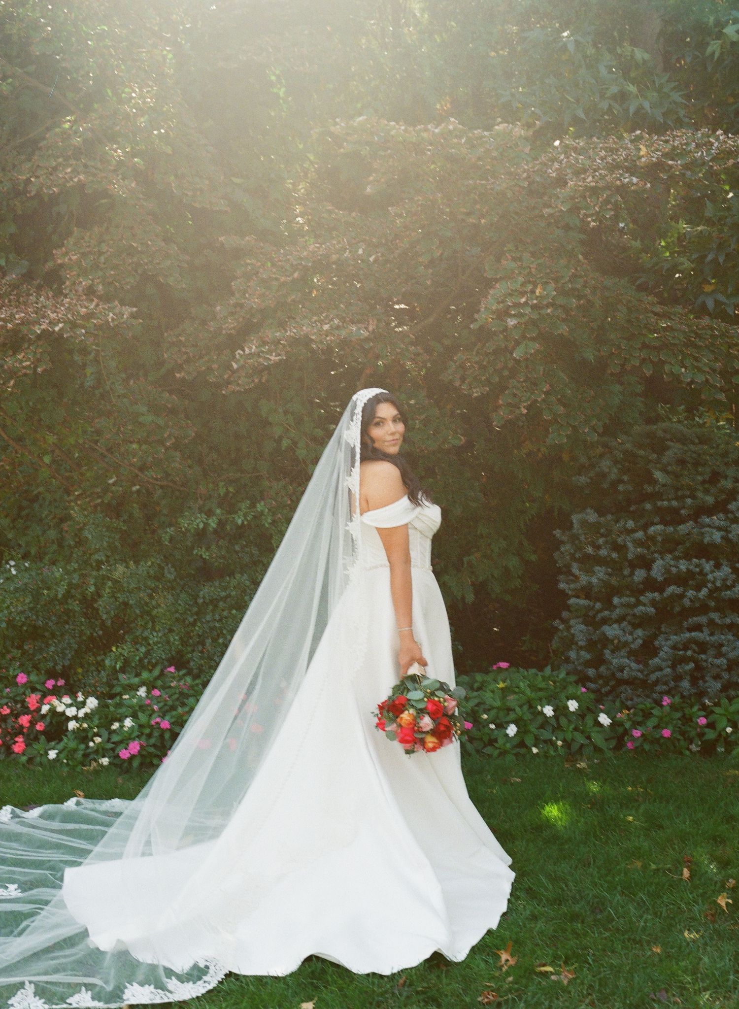 A bride in a flowing white wedding dress and veil poses in a sunlit garden setting with lush greenery.