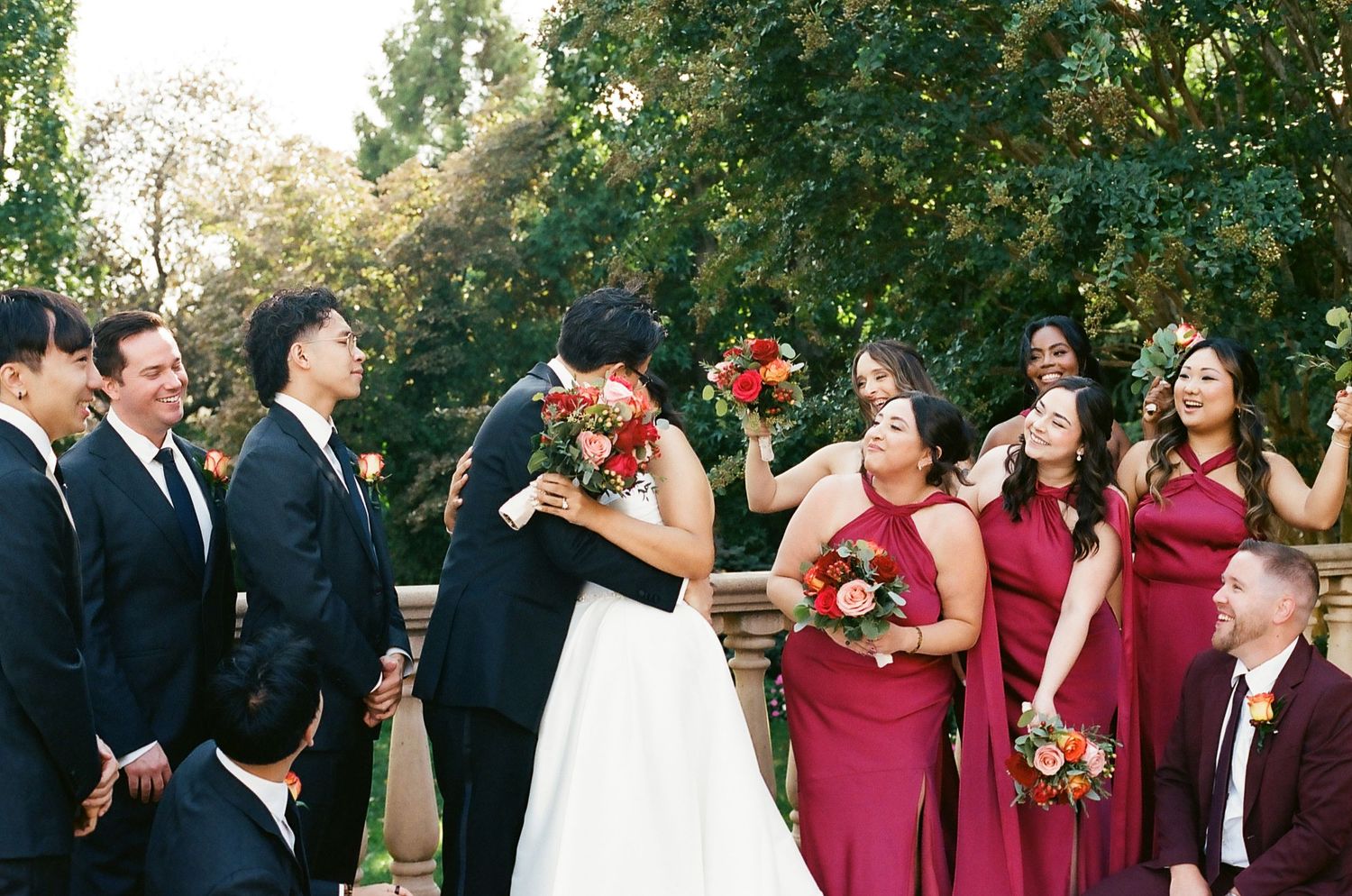 Wedding party in burgundy and black attire poses outdoors under trees during autumn celebration.