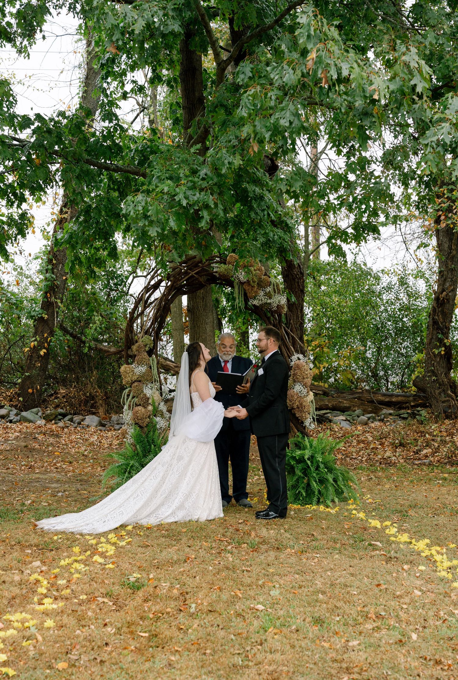 A wedding ceremony takes place under tall trees with scattered autumn leaves on the ground.
