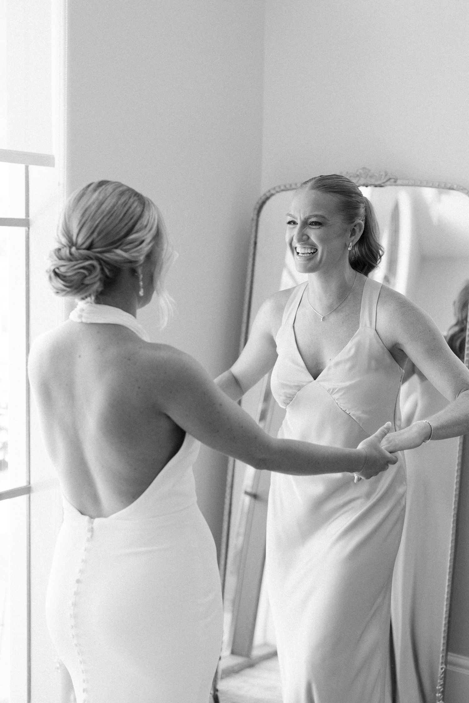 Two women in elegant white dresses share a joyful moment while getting ready in a bright room with a mirror.