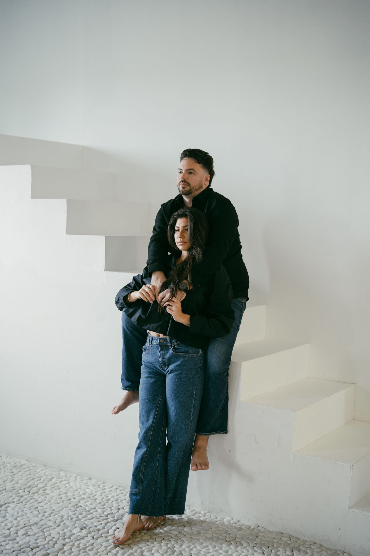 Two people in casual denim outfits stand barefoot on white stairs against a minimalist white wall in an artistic pose.