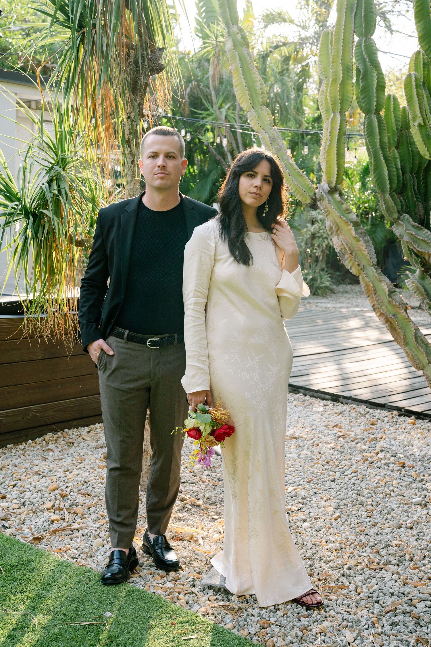 A couple poses together in a garden setting with tropical plants and gravel pathways.