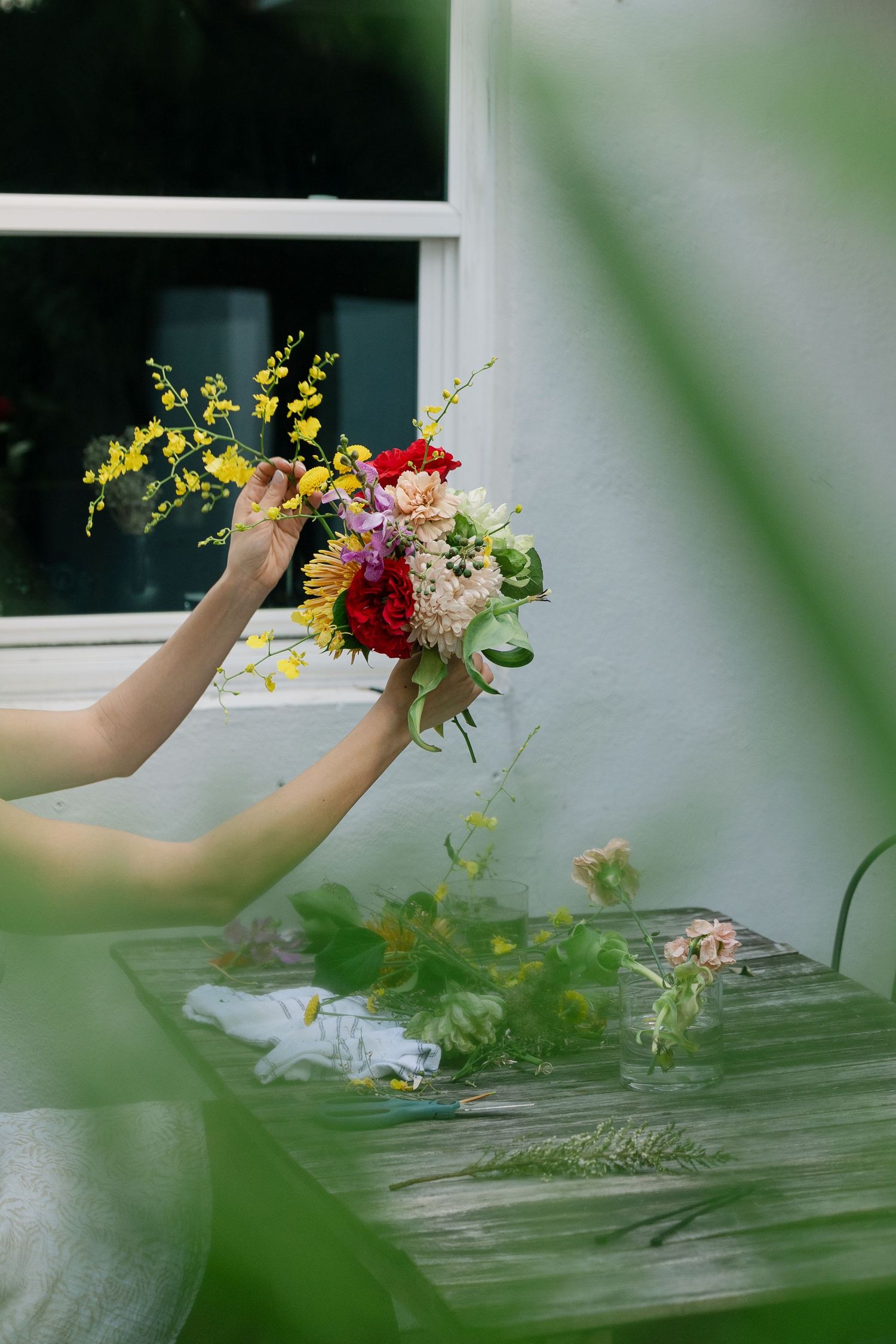 Hands arranging colorful fresh flowers in a rustic outdoor setting with natural greenery in the background.
