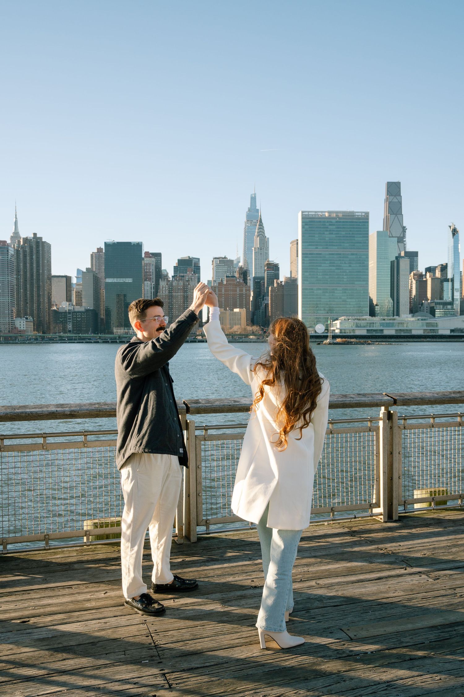 A couple dances playfully against the New York City skyline backdrop on a waterfront boardwalk.