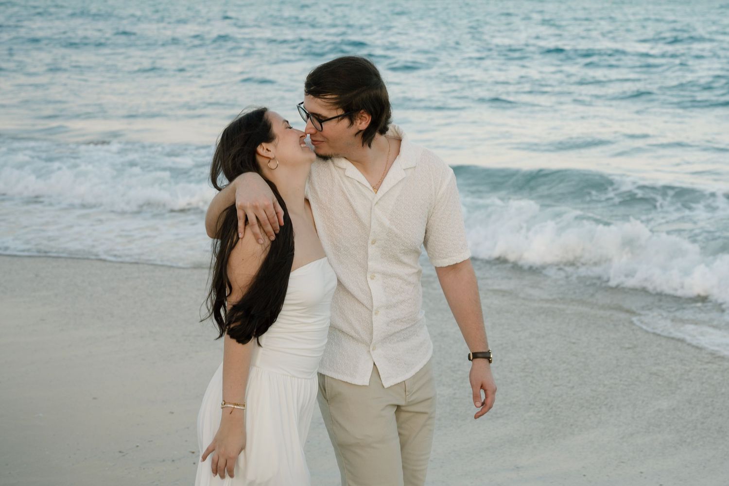 A couple shares a romantic moment walking along a sandy beach with ocean waves in the background.