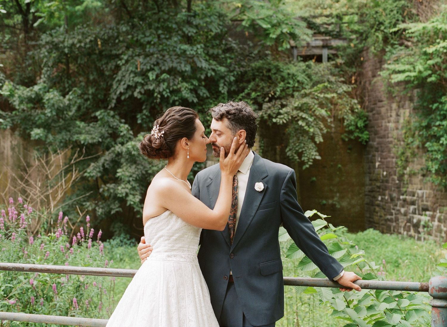 A couple shares an intimate moment in a lush garden setting with brick walls and green foliage surrounding them.
