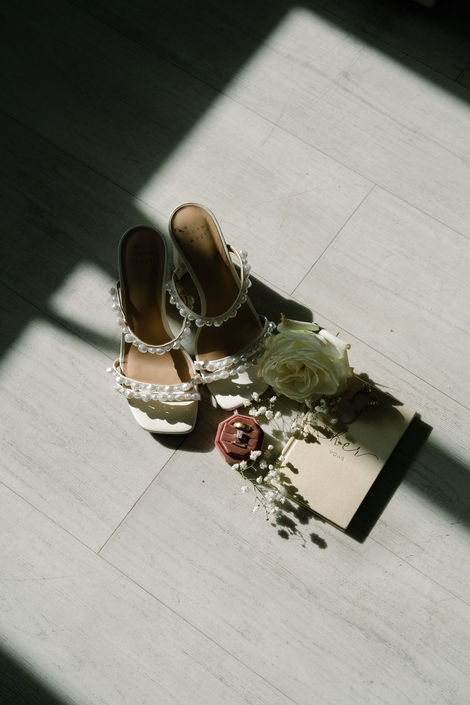 White bridal heels with pearl embellishments beside a rose, greenery, and an invitation card on a light wood floor.