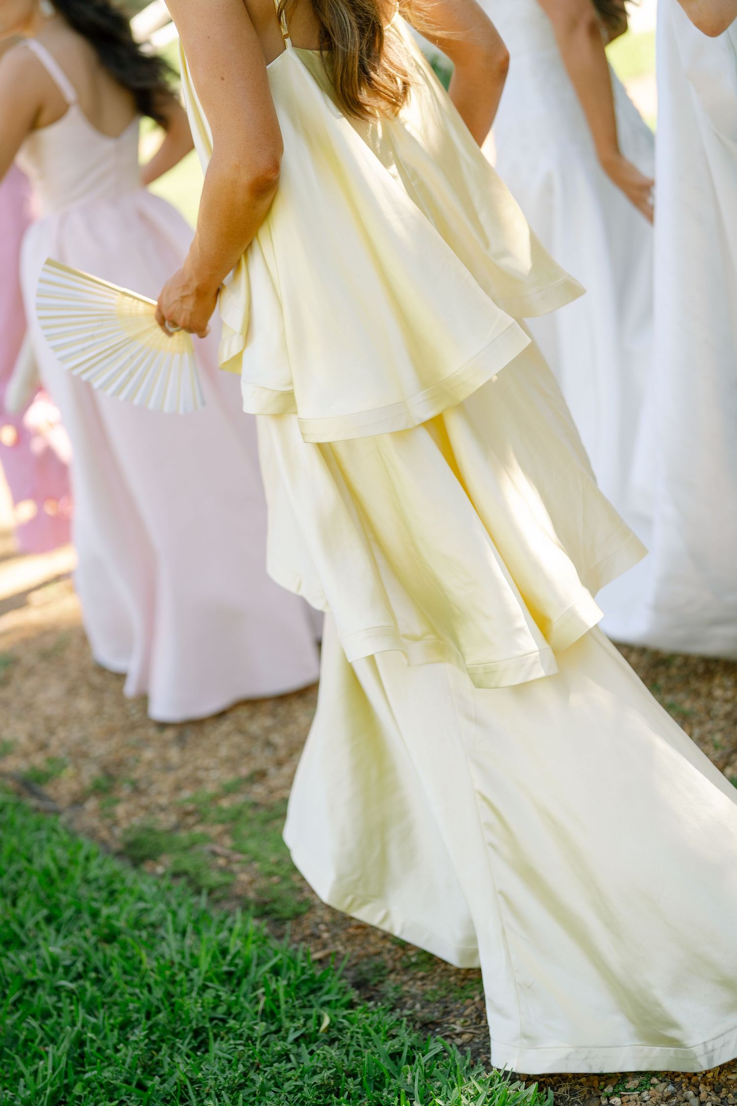 Bridesmaids in layered yellow and pink gowns holding decorative fans at an outdoor wedding.