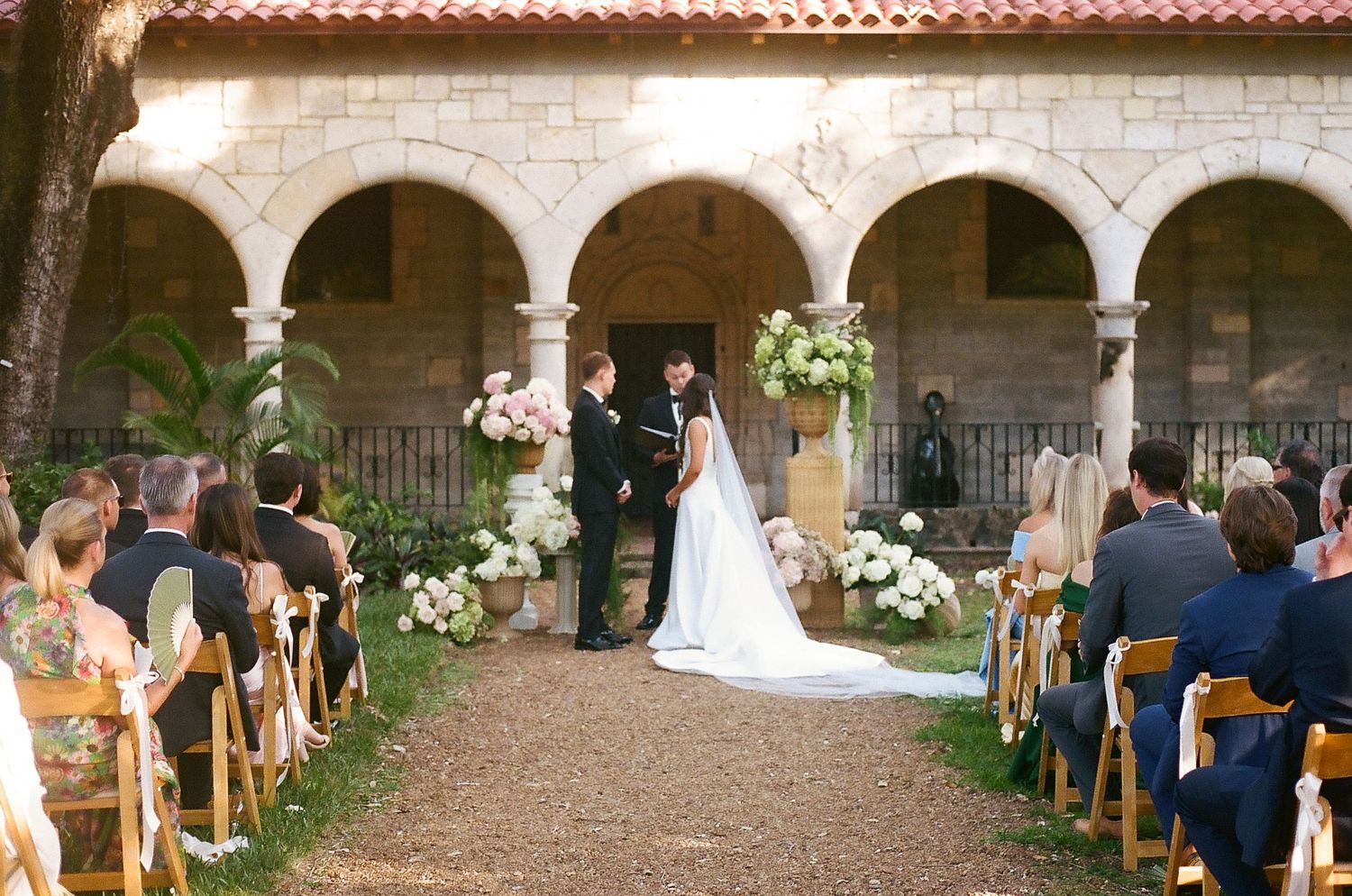 Elegant outdoor wedding ceremony in a courtyard with arched columns, floral arrangements, and guests seated on wooden chairs.