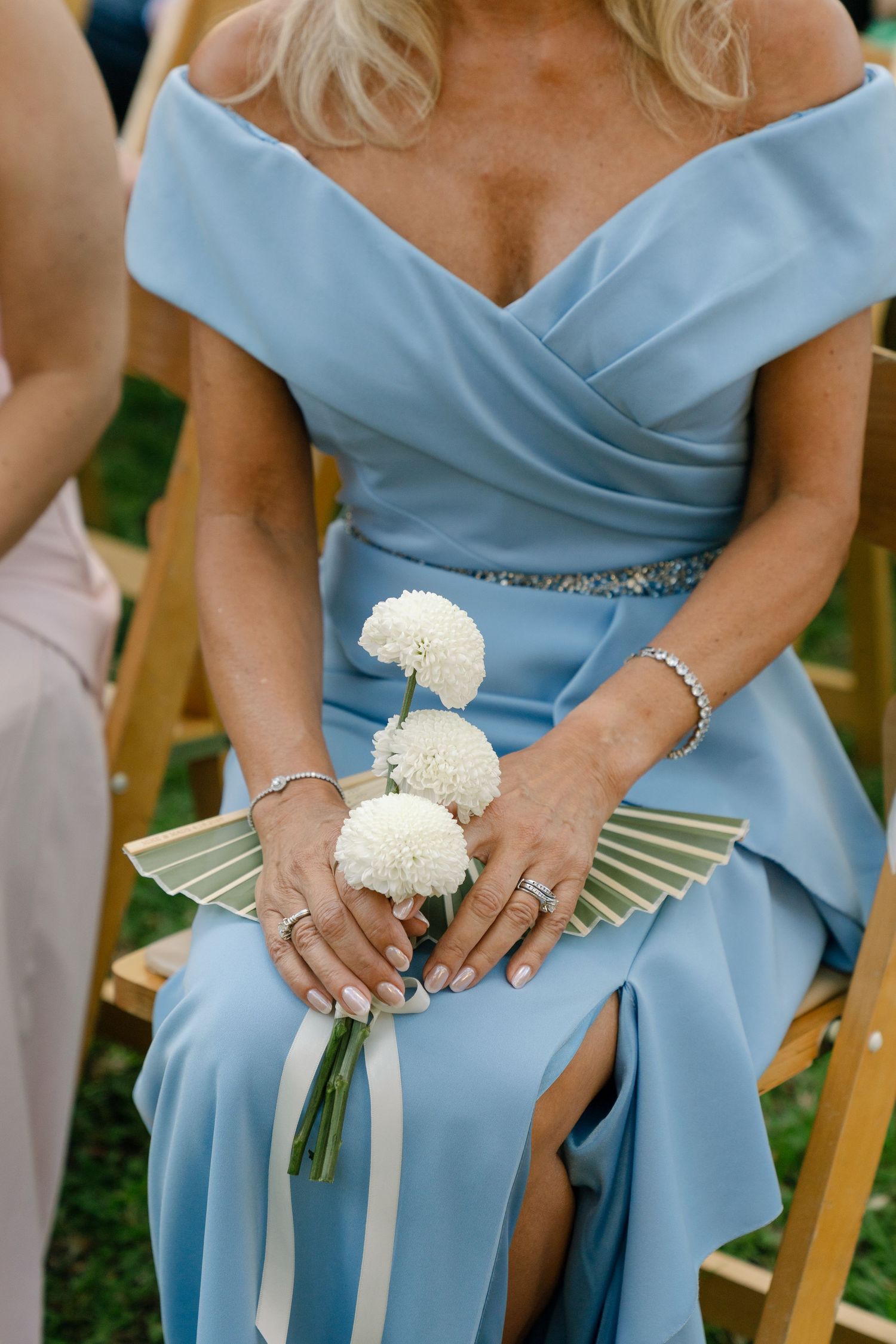 Woman in blue off-shoulder gown holds white pompom flowers and a fan at an outdoor wedding ceremony.