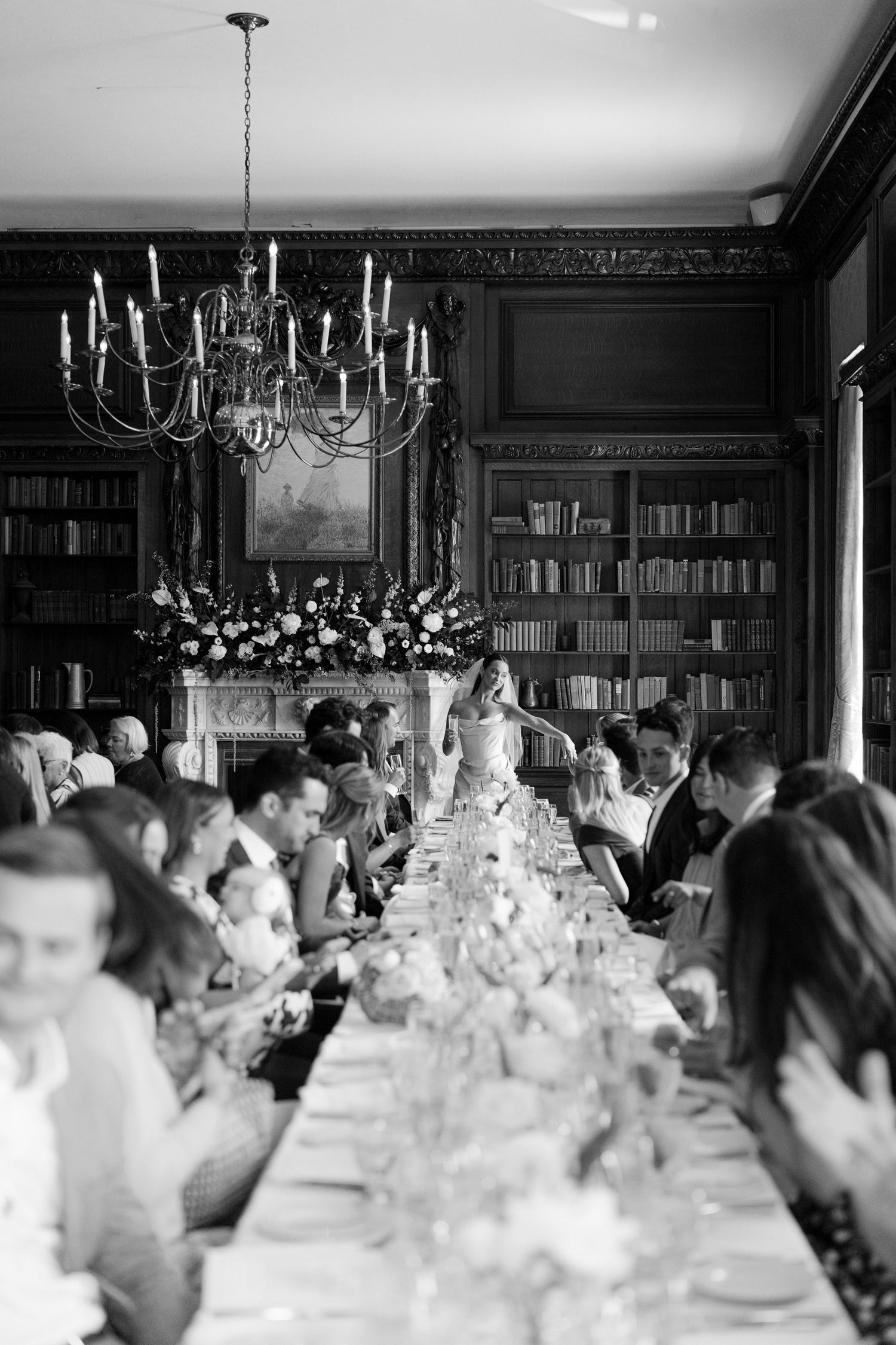 Elegant black and white wedding reception in a library dining room with chandelier and long table of guests.