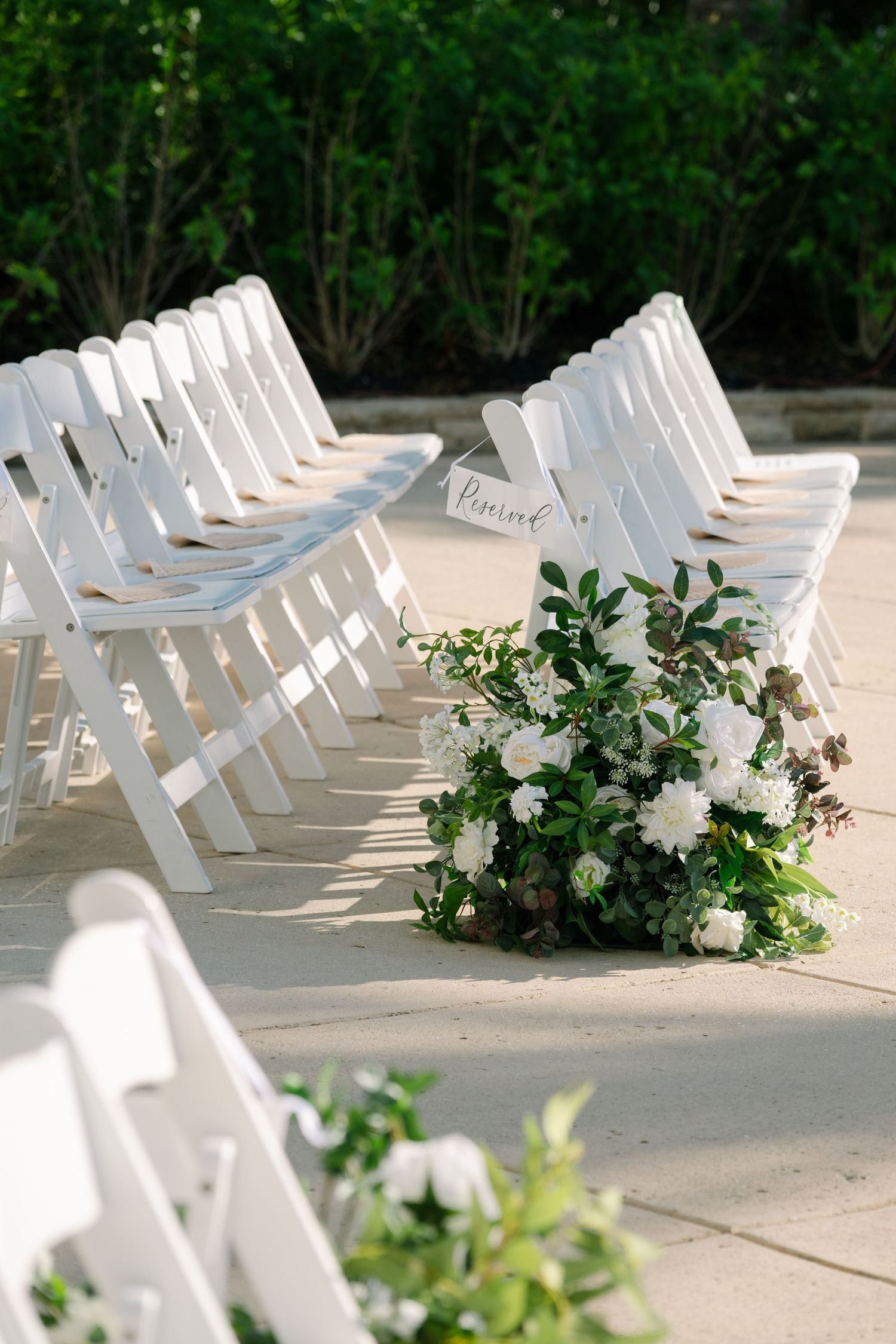 Row of white wedding ceremony chairs decorated with fresh floral arrangements on a sunny patio.