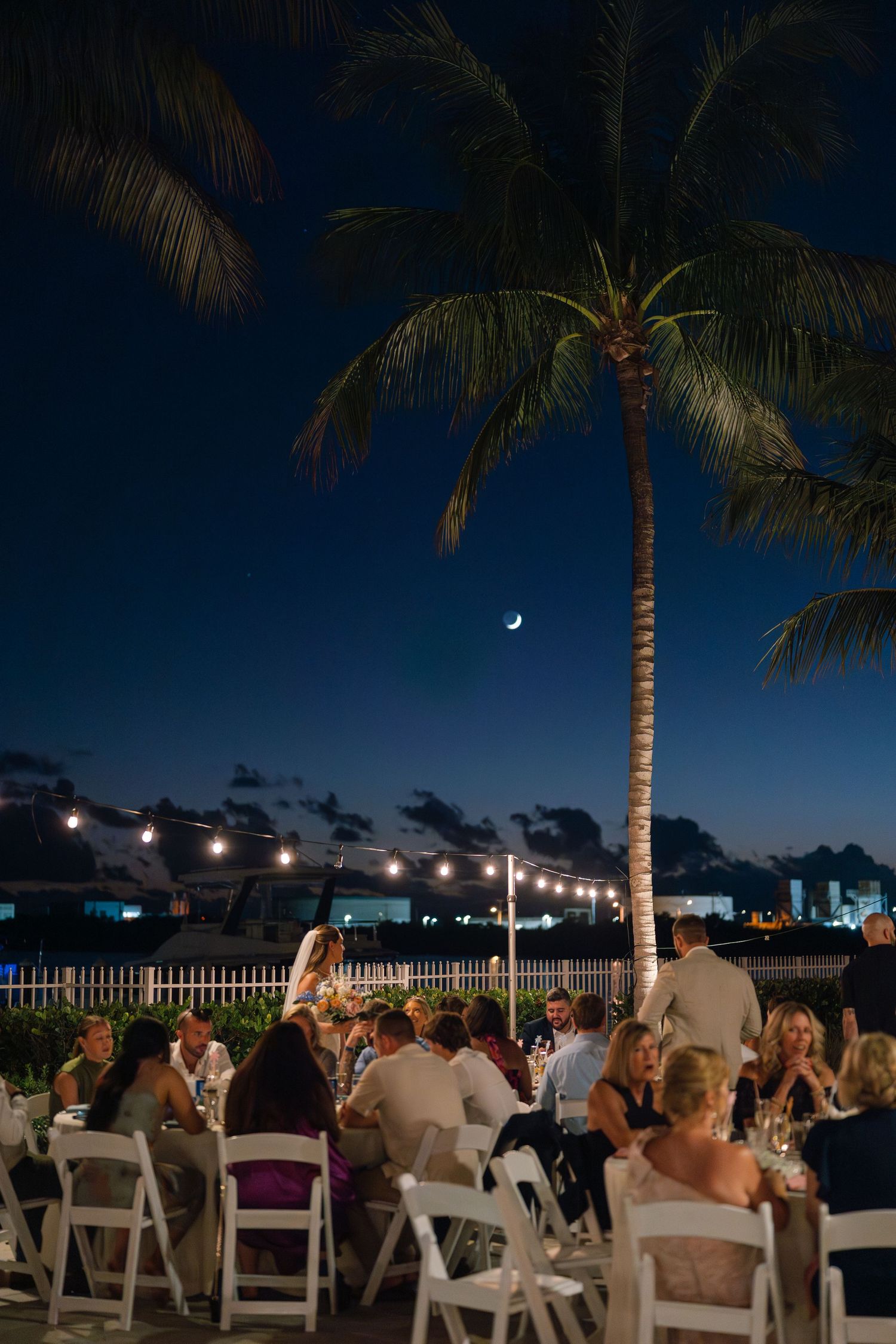Evening wedding reception under palm trees with string lights and crescent moon.