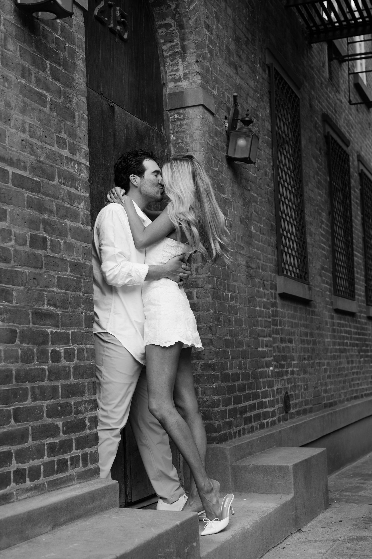 Black and white photo of a romantic couple kissing on steps against a brick wall building exterior.