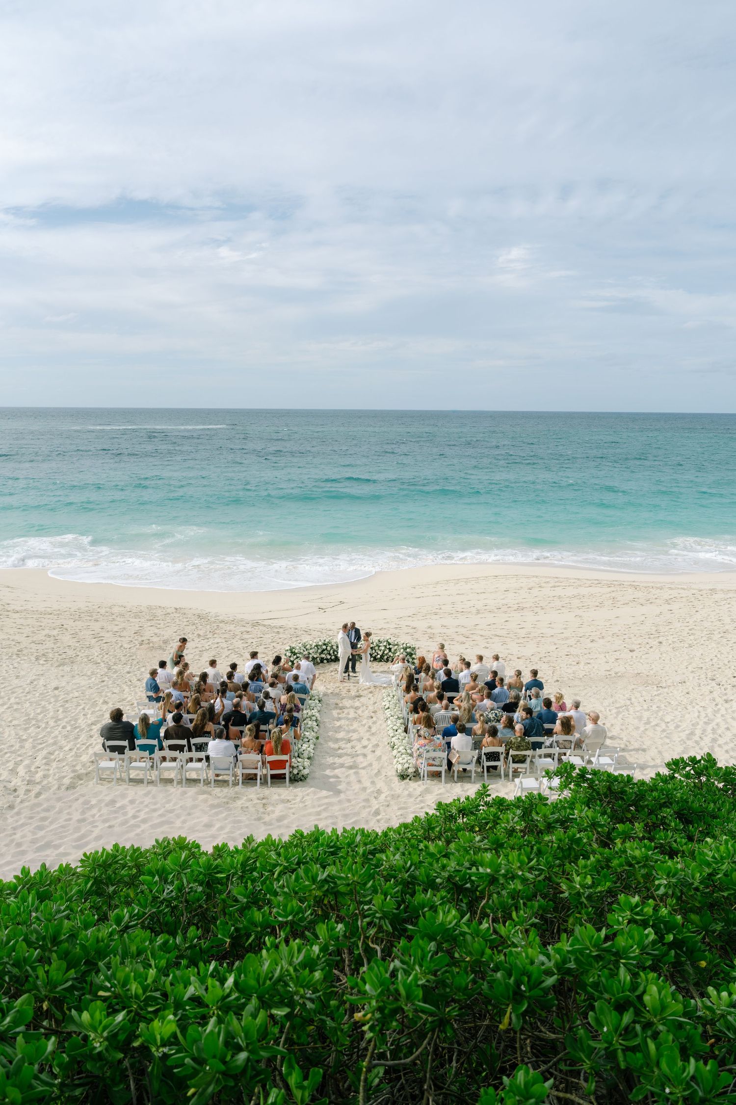 Aerial view of an intimate beach wedding ceremony with guests seated in white chairs facing the ocean on white sand.