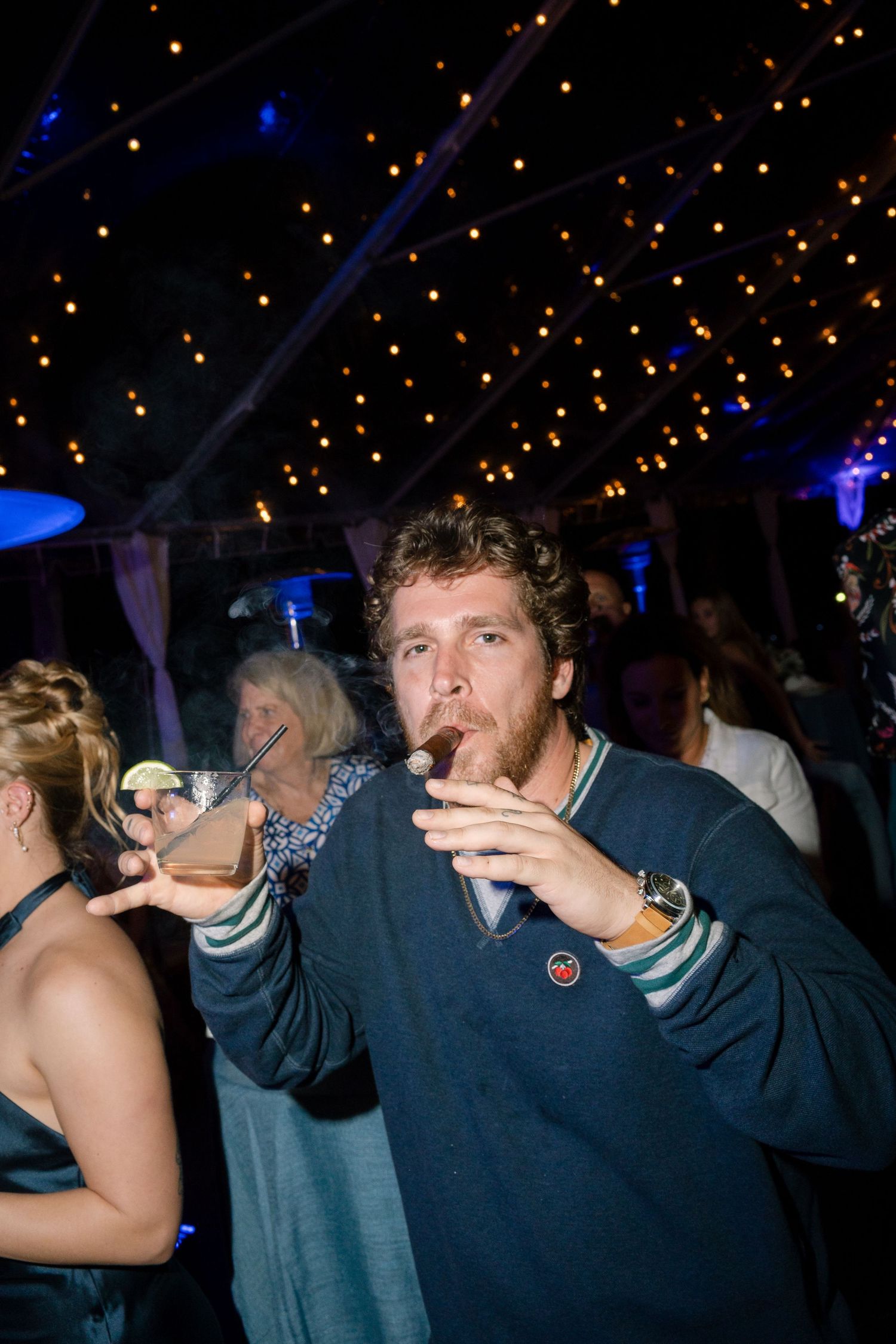Party guest enjoying a drink under decorative string lights at a nighttime celebration.