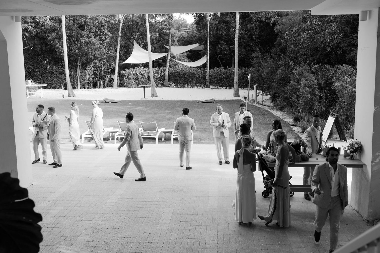 Wedding guests gather on an outdoor patio area with white sail-like canopies overhead.