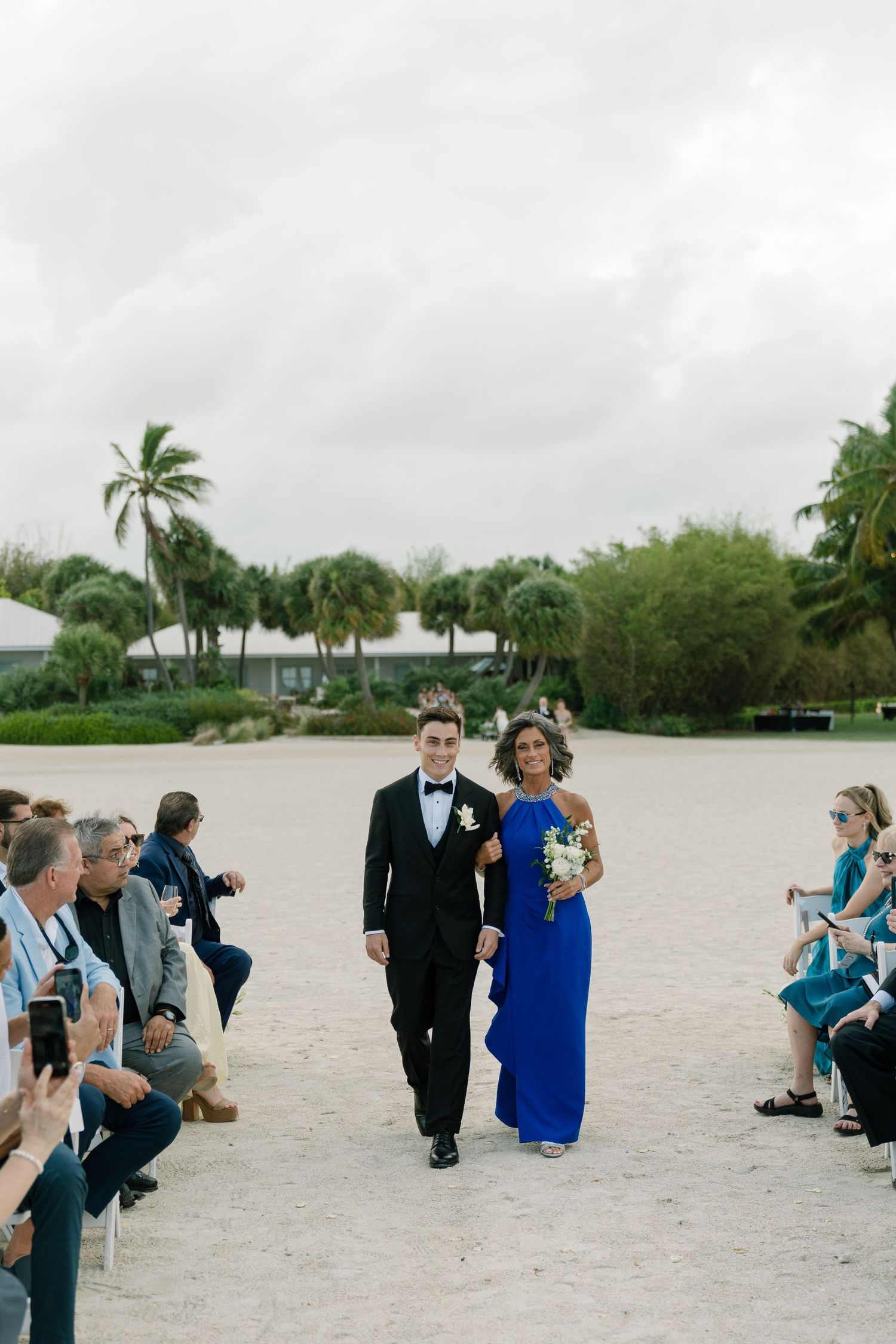 Groom in a black tuxedo walks down the sandy ceremony aisle escorted by a woman in a royal blue gown.