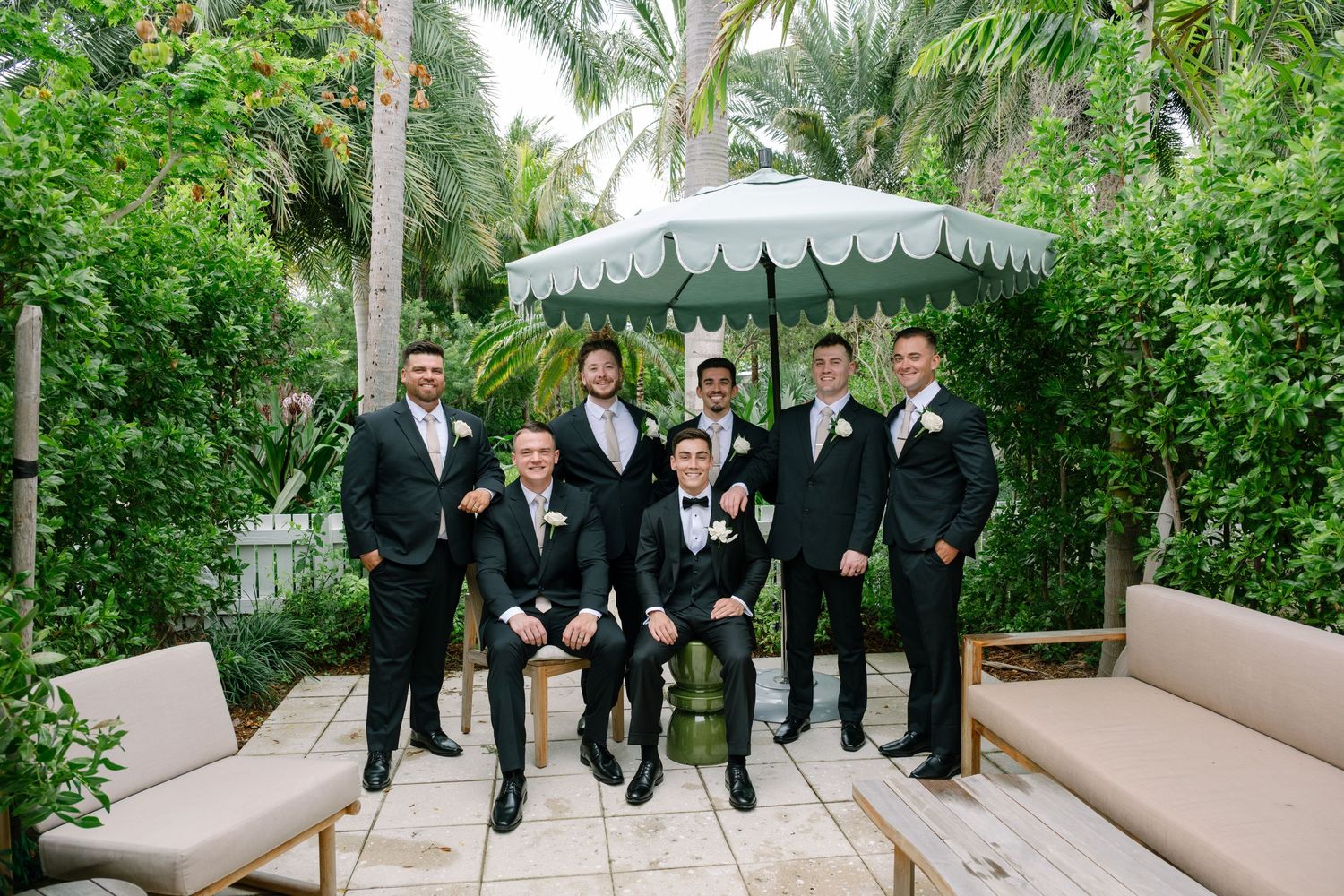 Groom poses with groomsmen in black tuxedos under white umbrella in tropical garden.