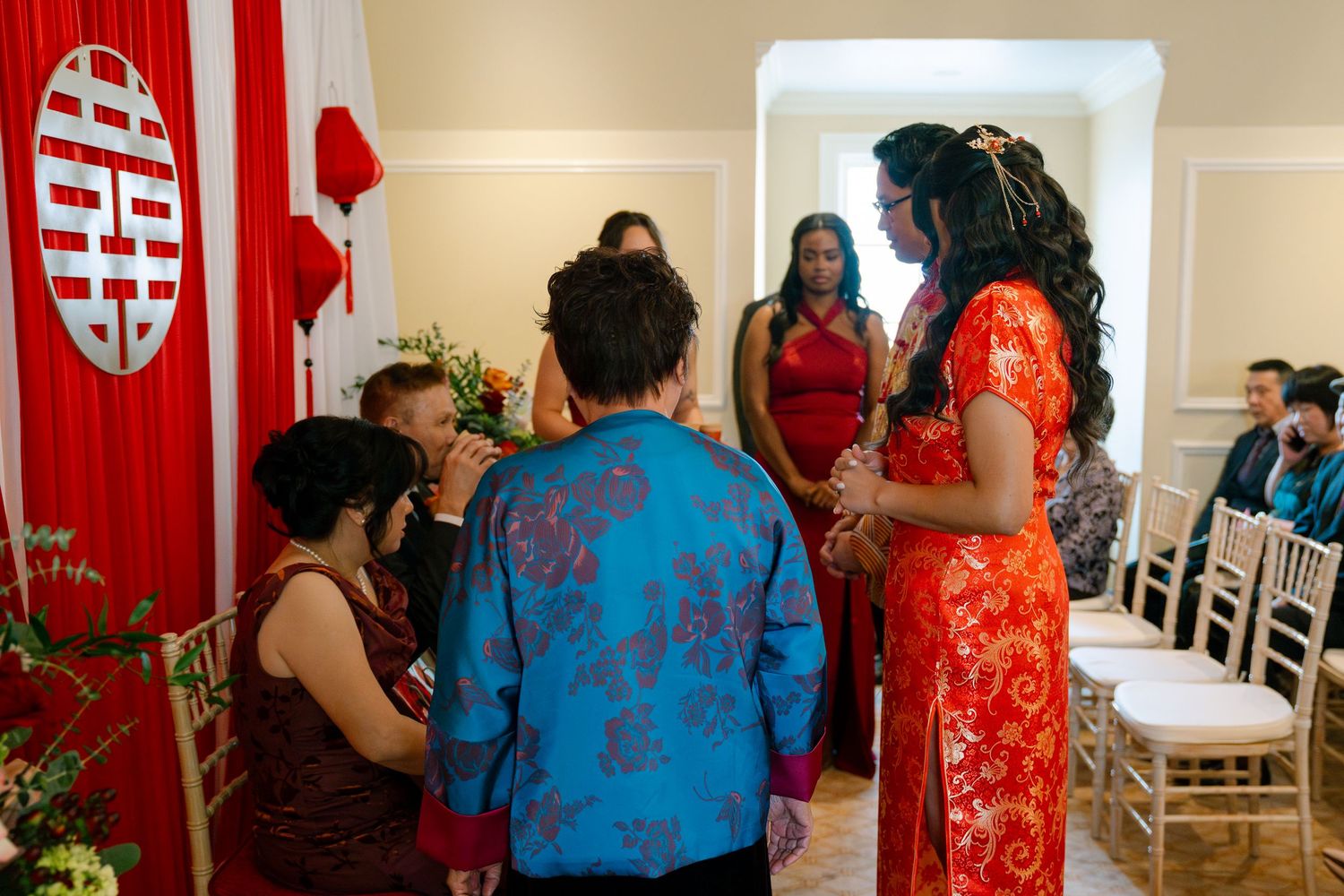 Wedding guests gather in a room decorated with red and white traditional Chinese elements and white chairs.