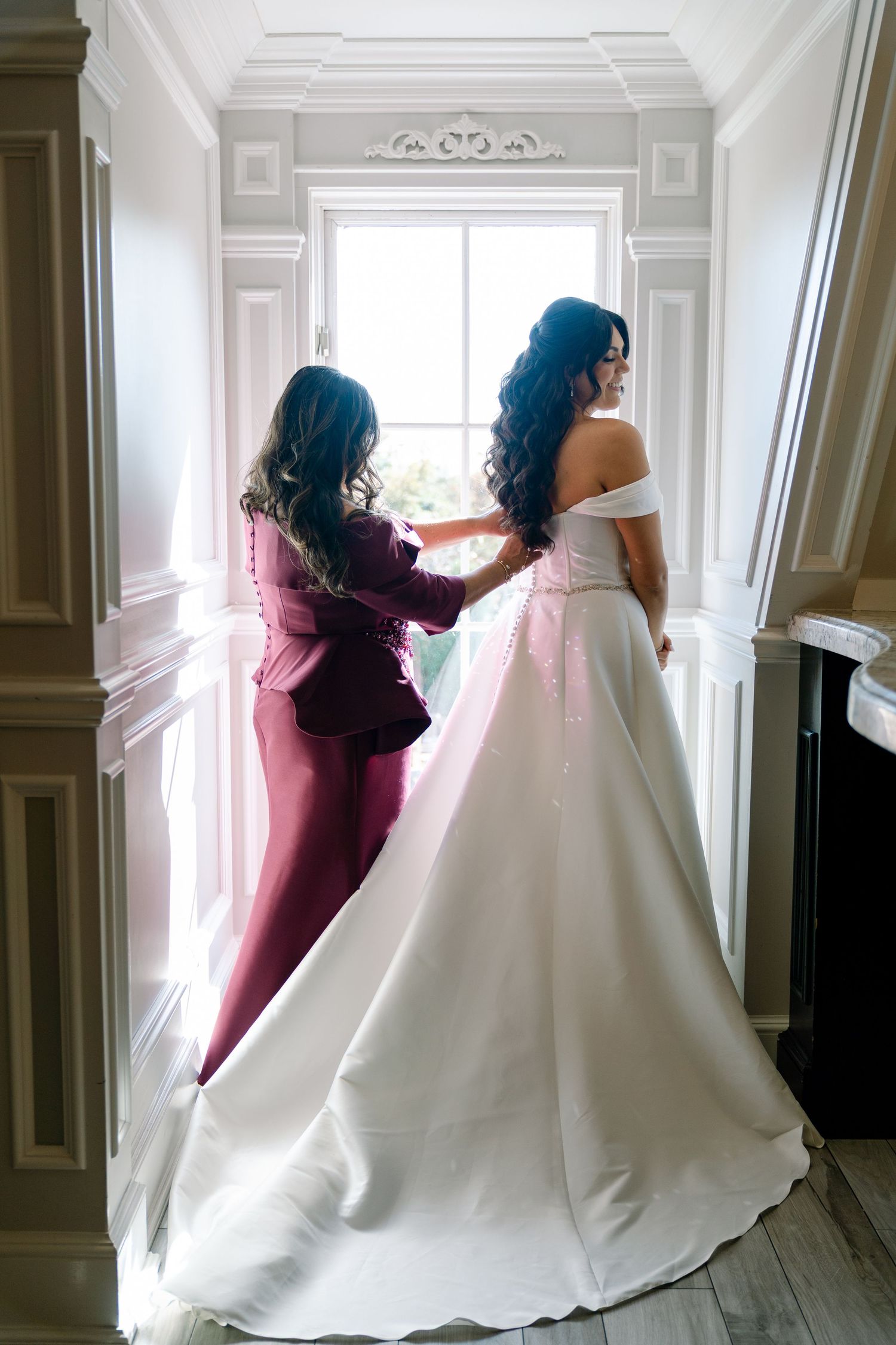 A helper adjusts the back of an elegant white wedding gown in a bright hallway with ornate molding.