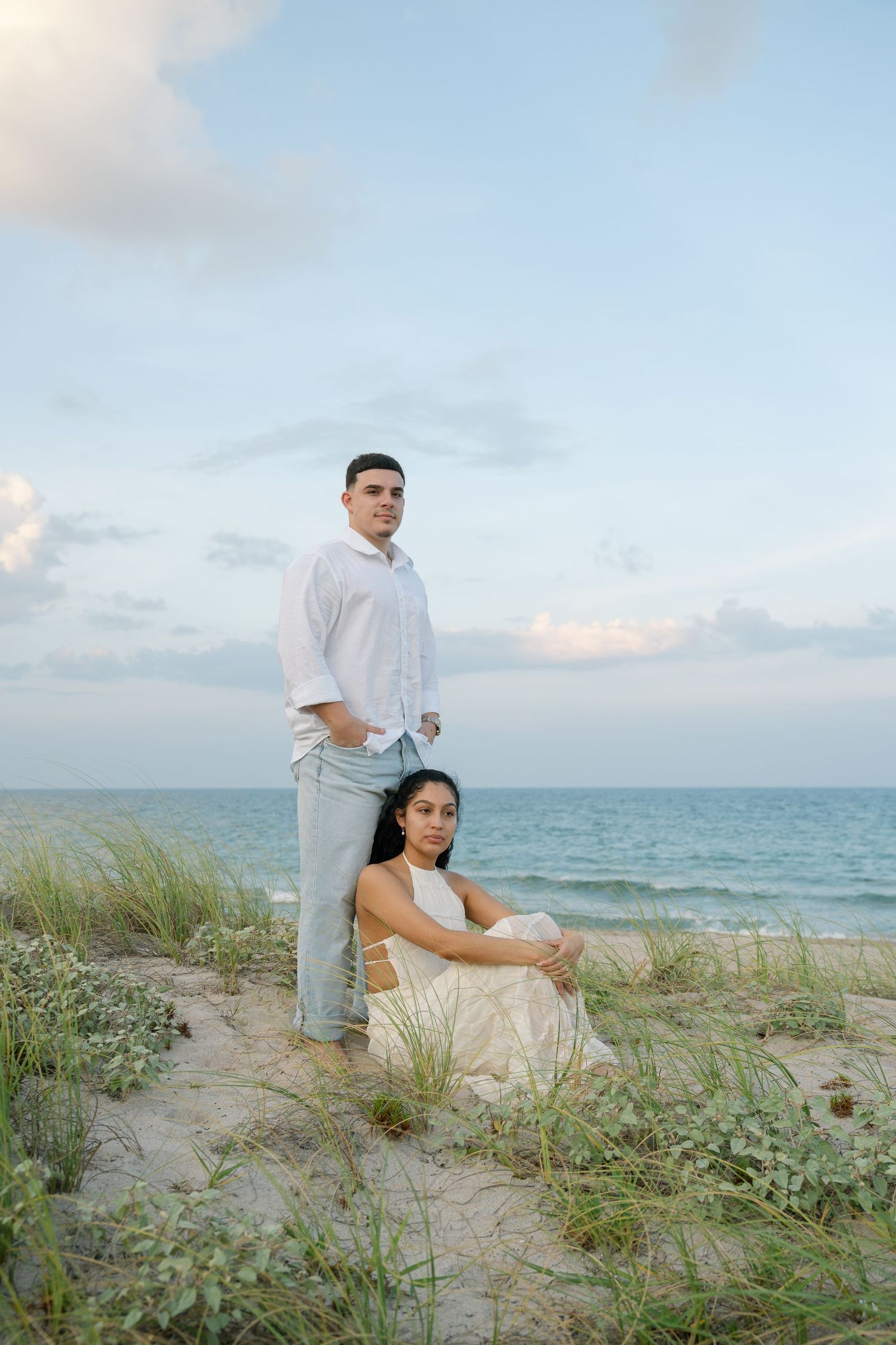 A couple poses together on sandy dunes with beach grass and ocean views at sunset.