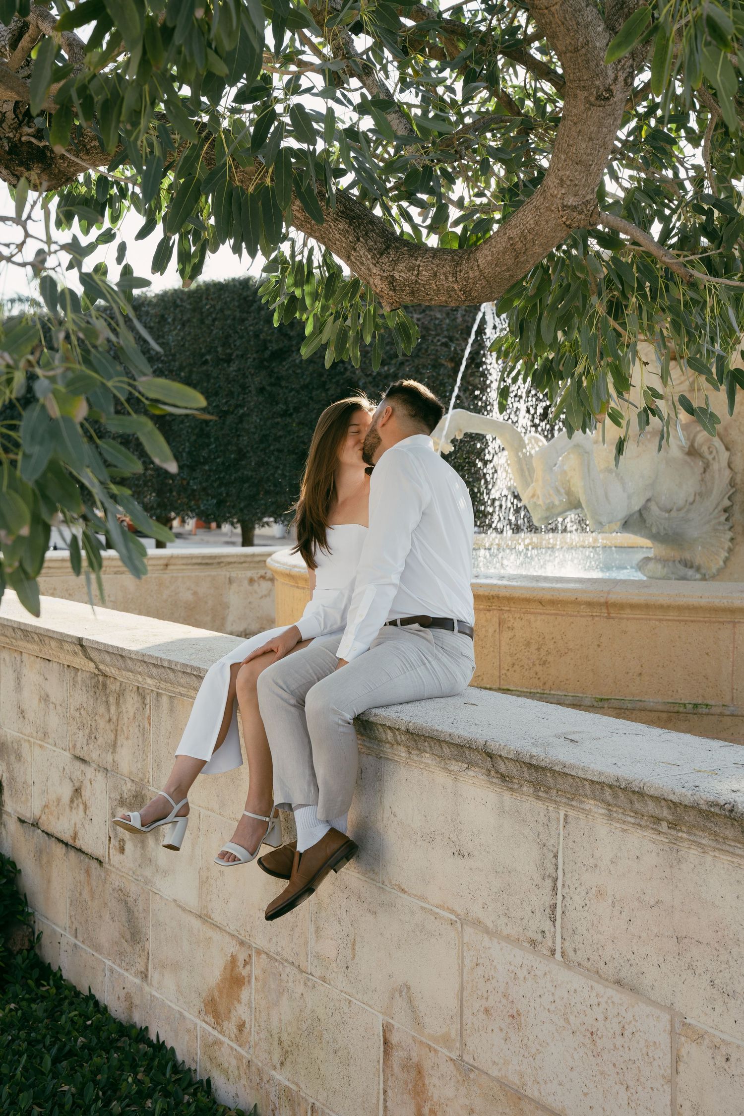 A person in a white outfit sits casually on a stone wall under a tree with green leaves overhead.