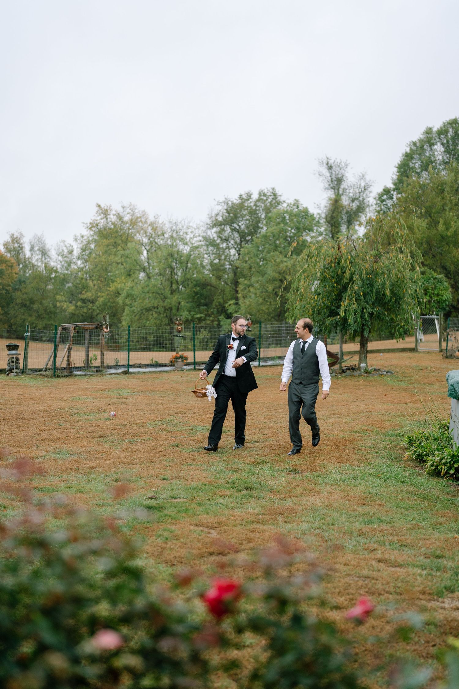 Two people in formal attire walk through an open field with trees in the background.