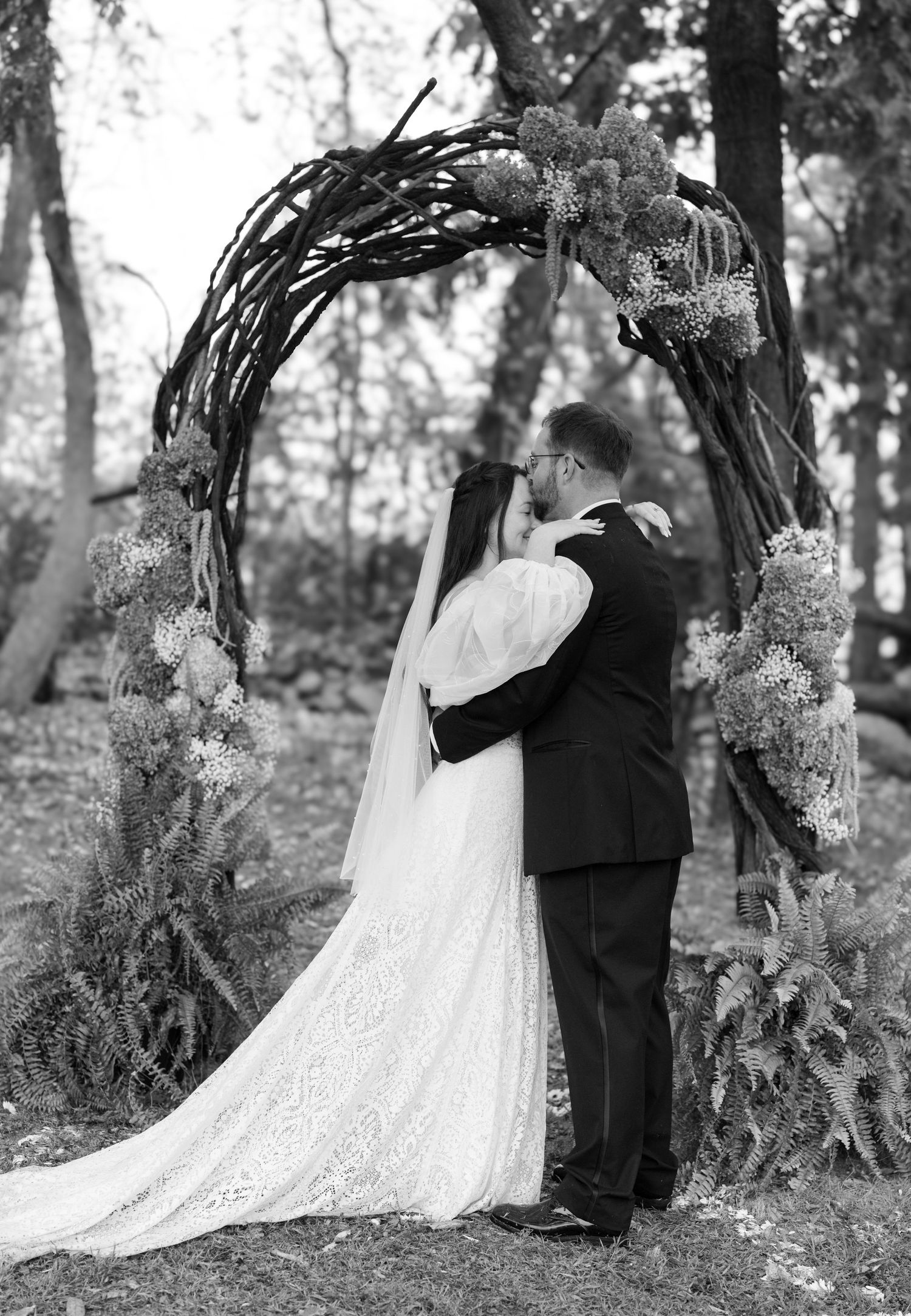 A romantic wedding couple embraces under a rustic wooden arch decorated with delicate floral arrangements in a forest setting.