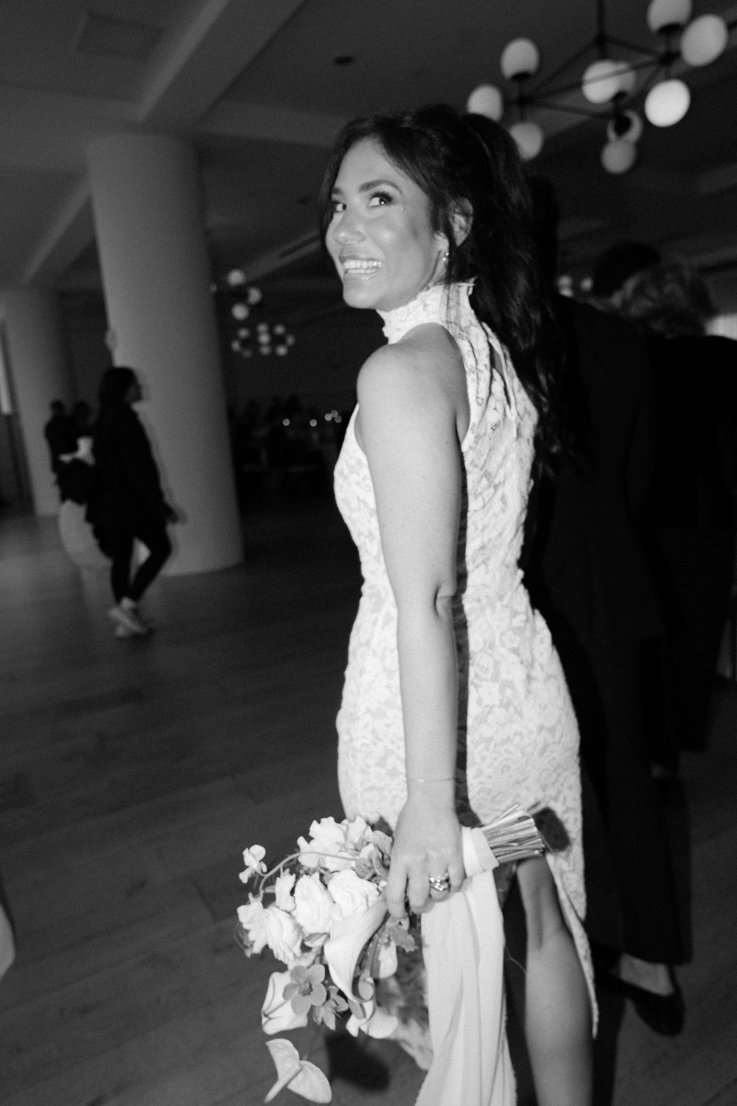 A bride in a white lace dress holds a bouquet while standing in an indoor venue with overhead lighting.