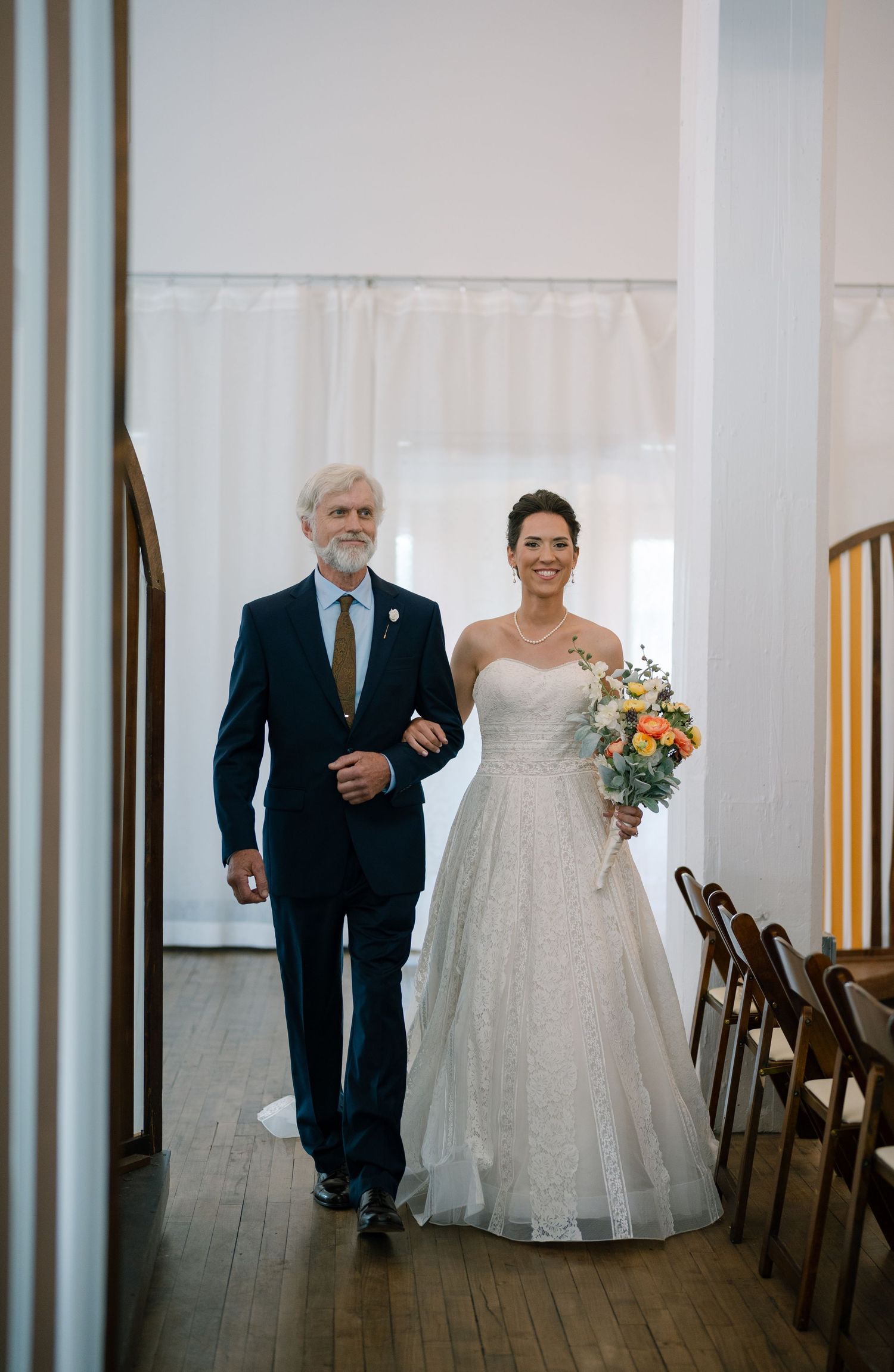 A wedding party walks down a hallway with wooden floors and white walls.