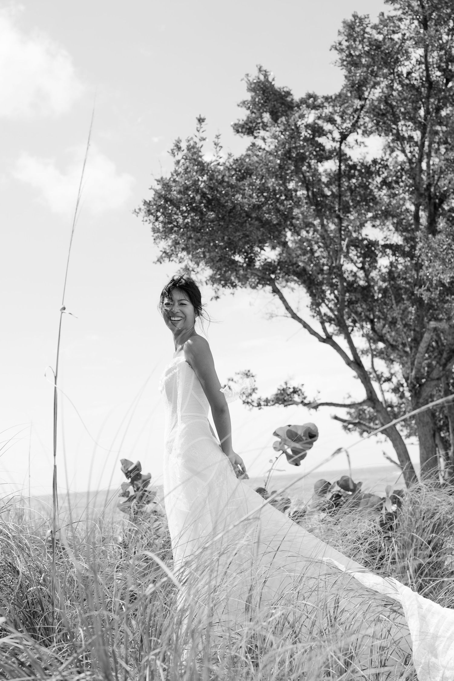 Smiling bride in white gown stands among tall grass with trees and sky in black and white outdoor photo.