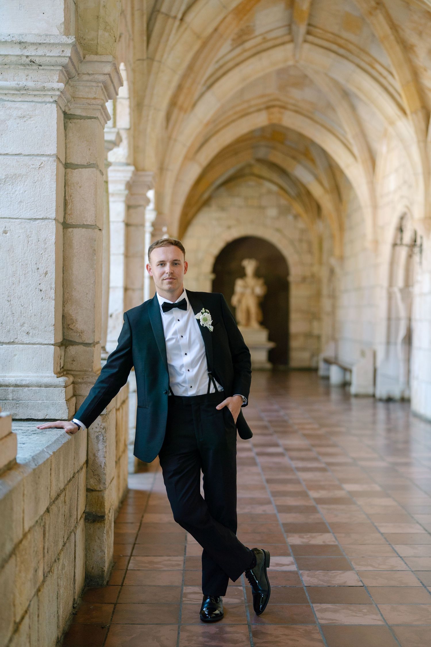 Groom in black tuxedo leans against column in elegant arched stone corridor with marble floors.