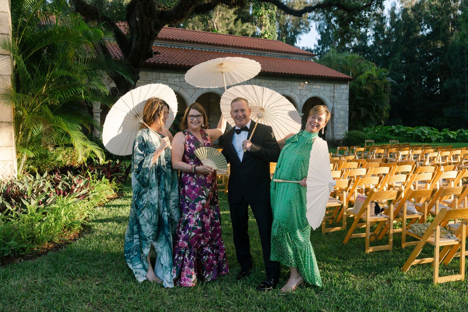 Wedding guests holding white parasols pose outside stone chapel at outdoor ceremony venue.