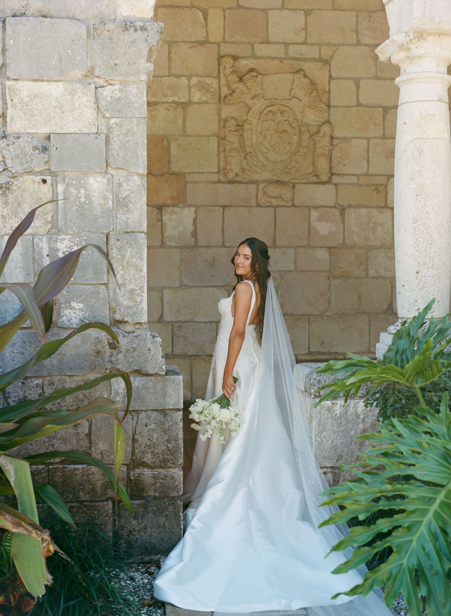 Bride in elegant white gown with veil holds bouquet beside ornate stone arch surrounded by tropical greenery.