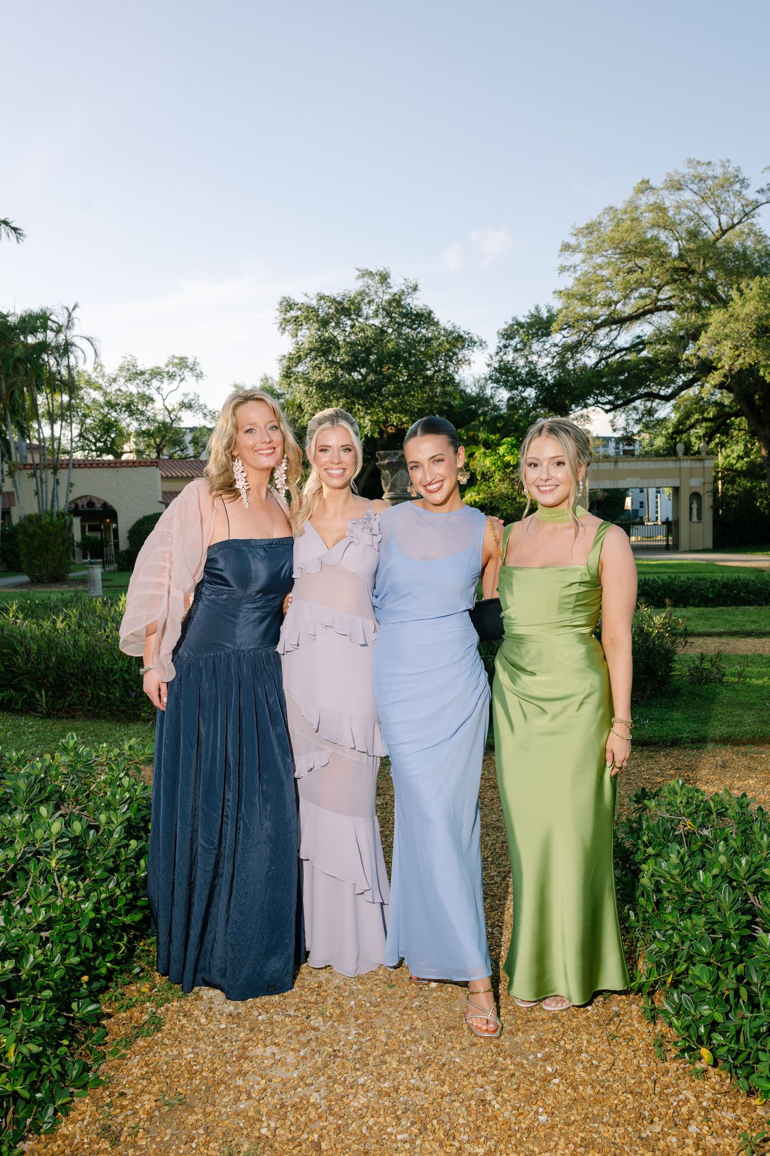 Four women in elegant mismatched evening gowns pose together smiling in a manicured garden at wedding reception.