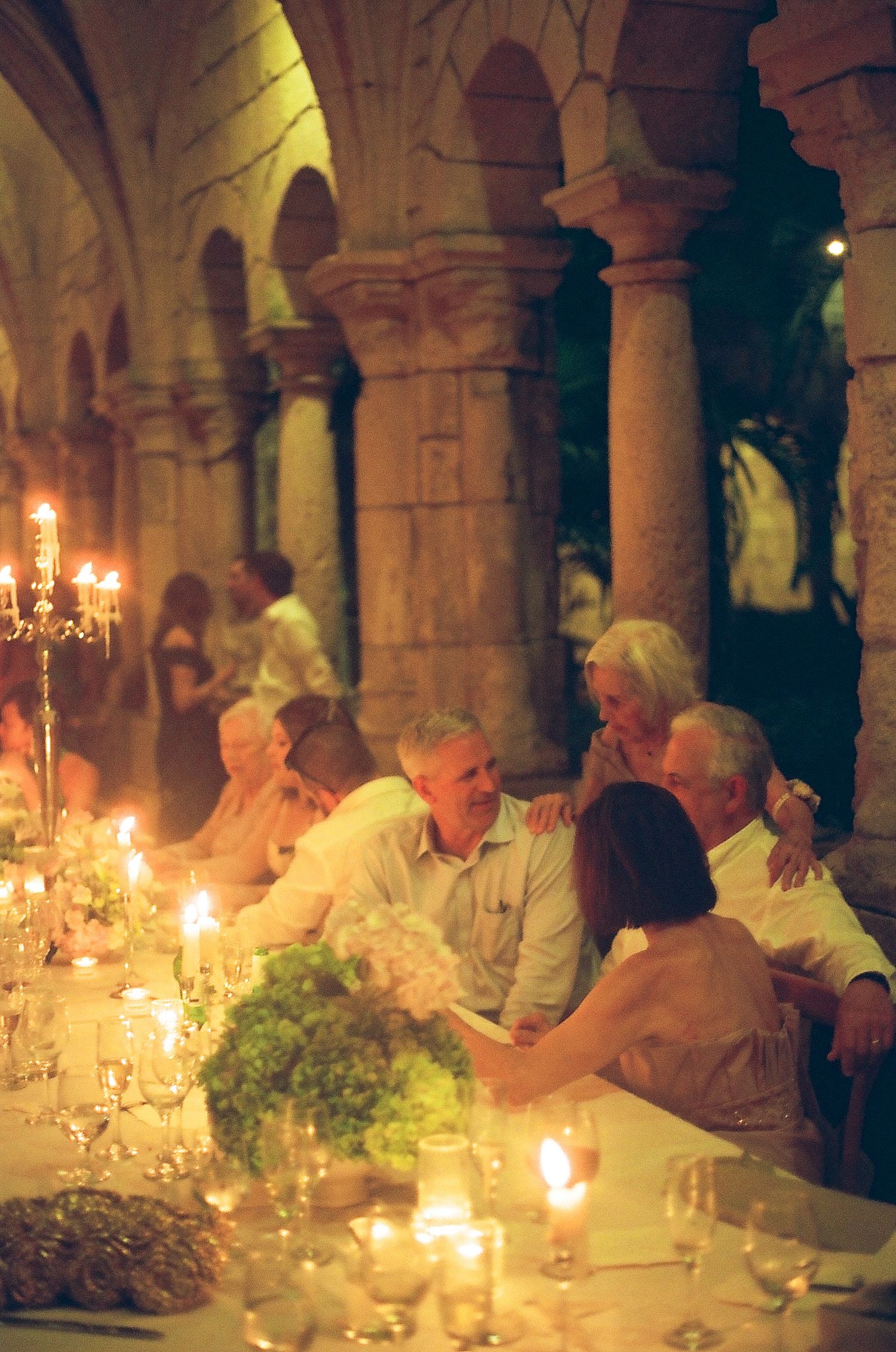 Guests enjoy a candlelit wedding reception dinner beneath stone arches and columns in an elegant venue.